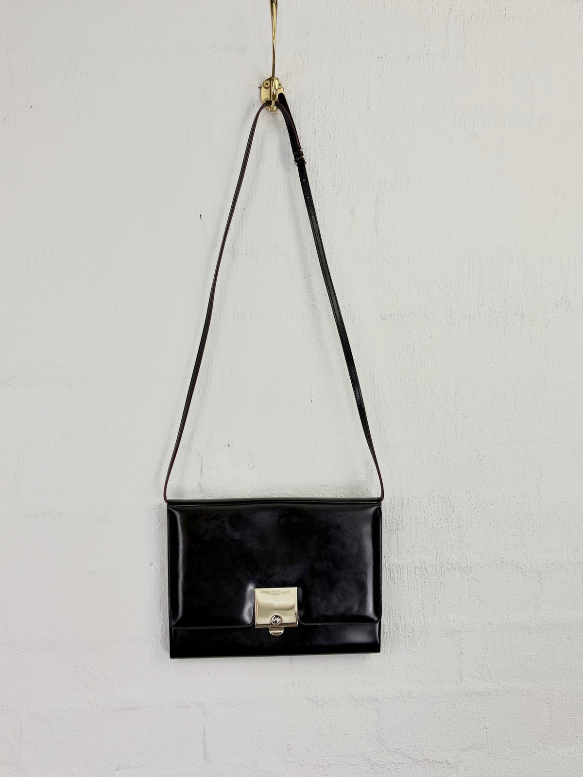 A black glossy shoulder bag with a rectangular silhouette and a gold-tone clasp, hanging from a gold hook against a white textured wall.