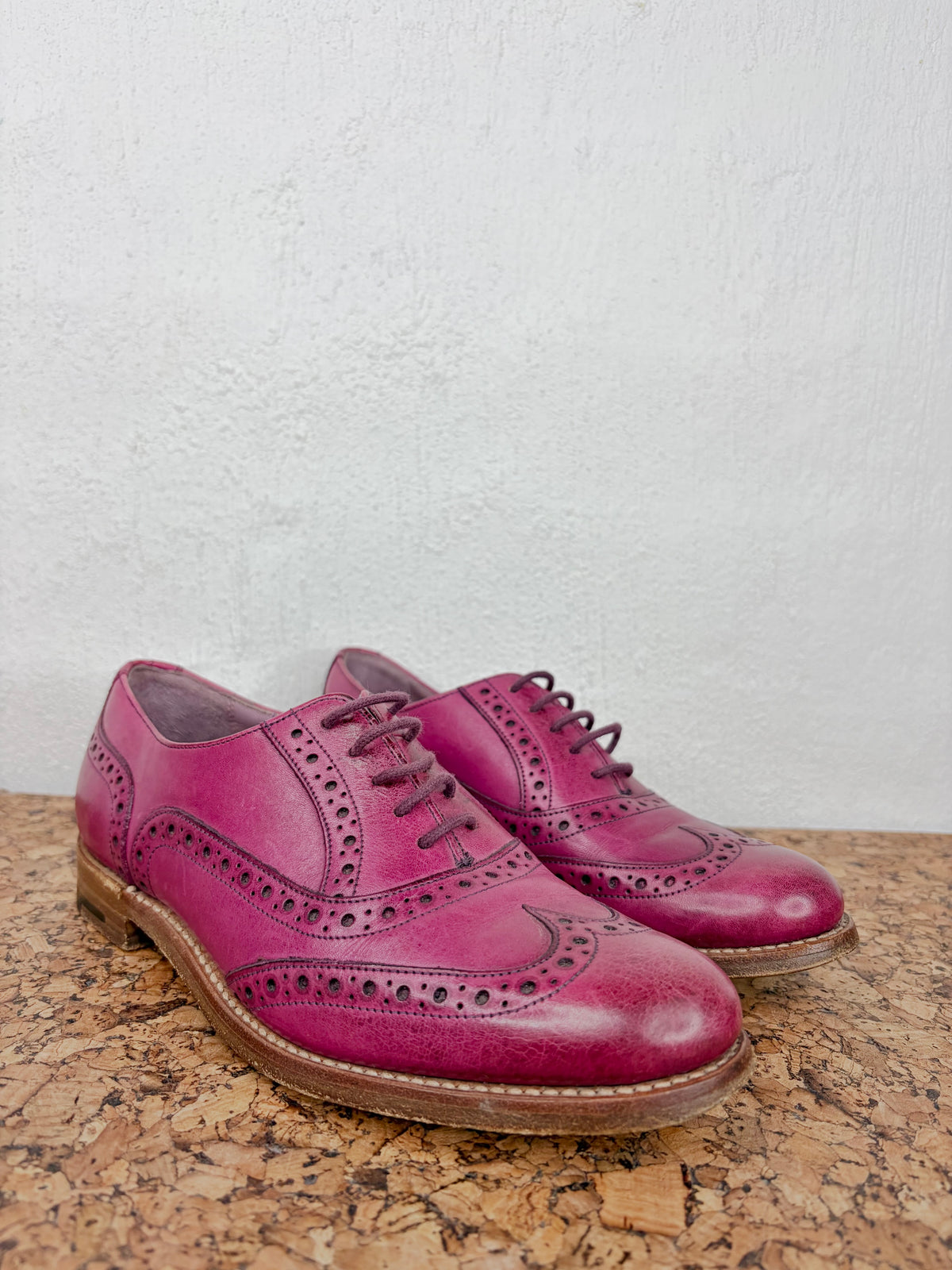 A pair of magenta leather wingtip oxford shoes with decorative brogue perforations and brown laces, resting on a cork surface against a textured white wall.