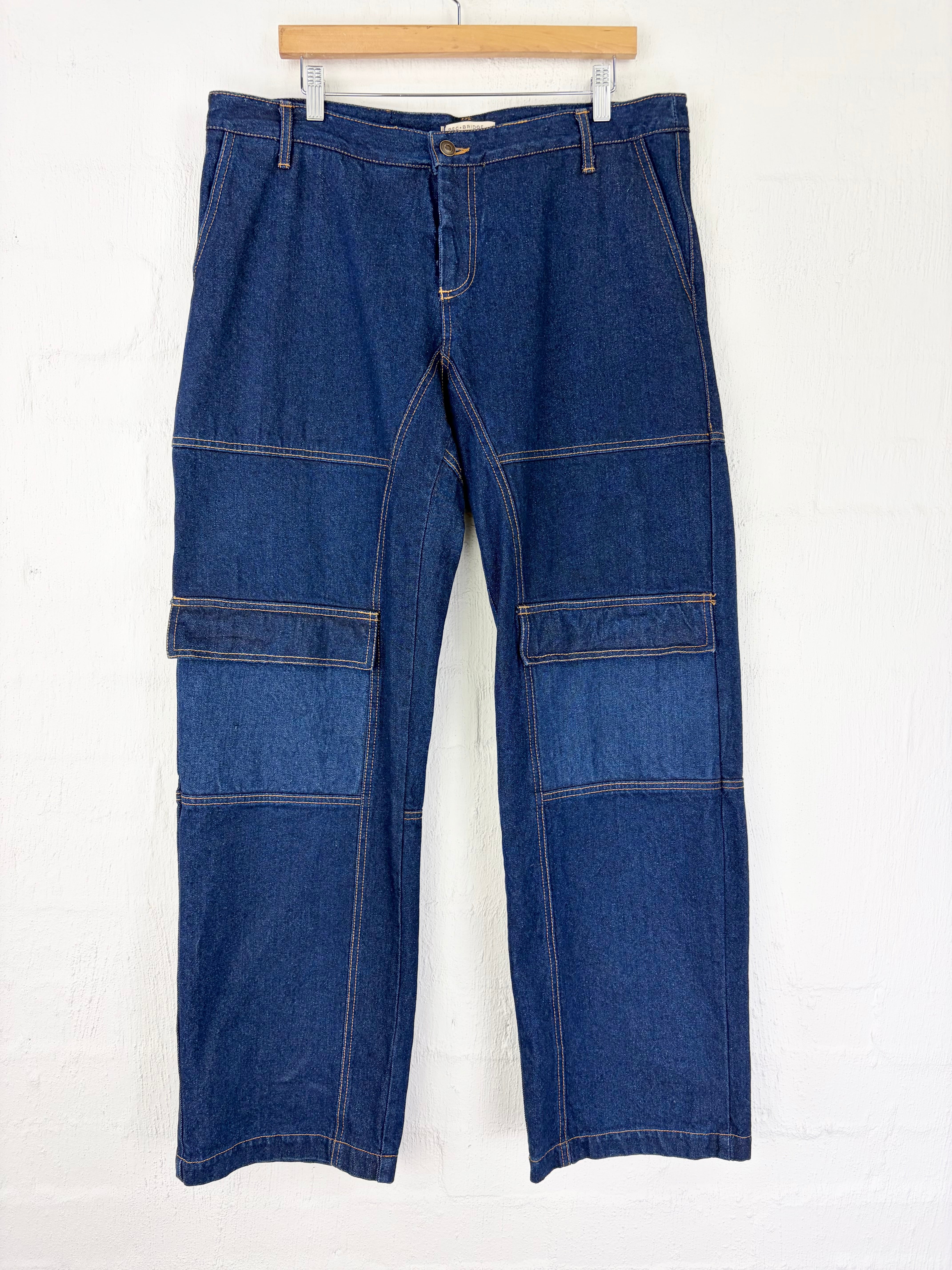 A pair of dark blue denim pants with patchwork paneling and contrast tan stitching, hanging on a wooden hanger against a white wall.