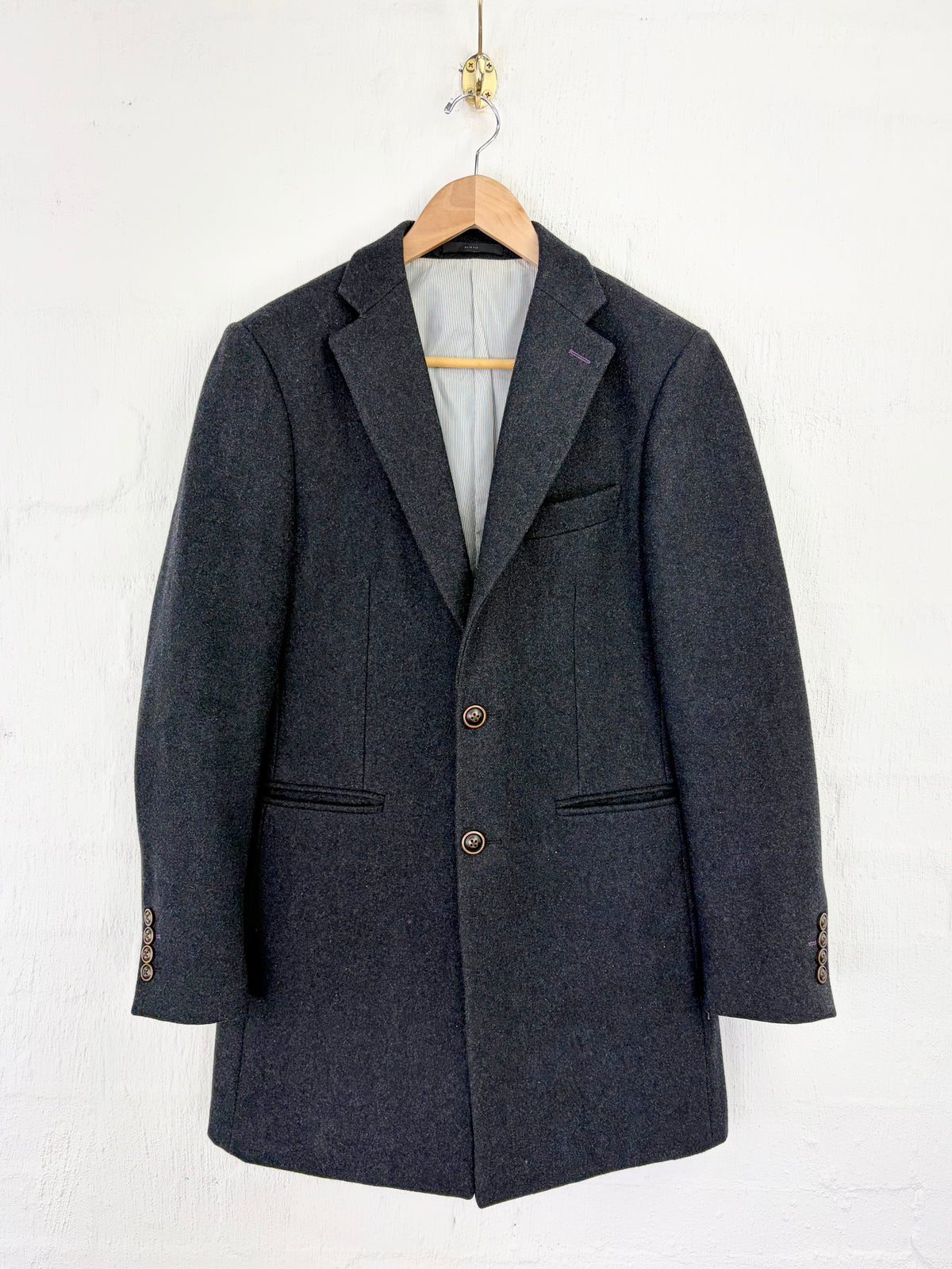 A dark gray textured wool blazer hanging on a wooden hanger against a white painted brick wall.