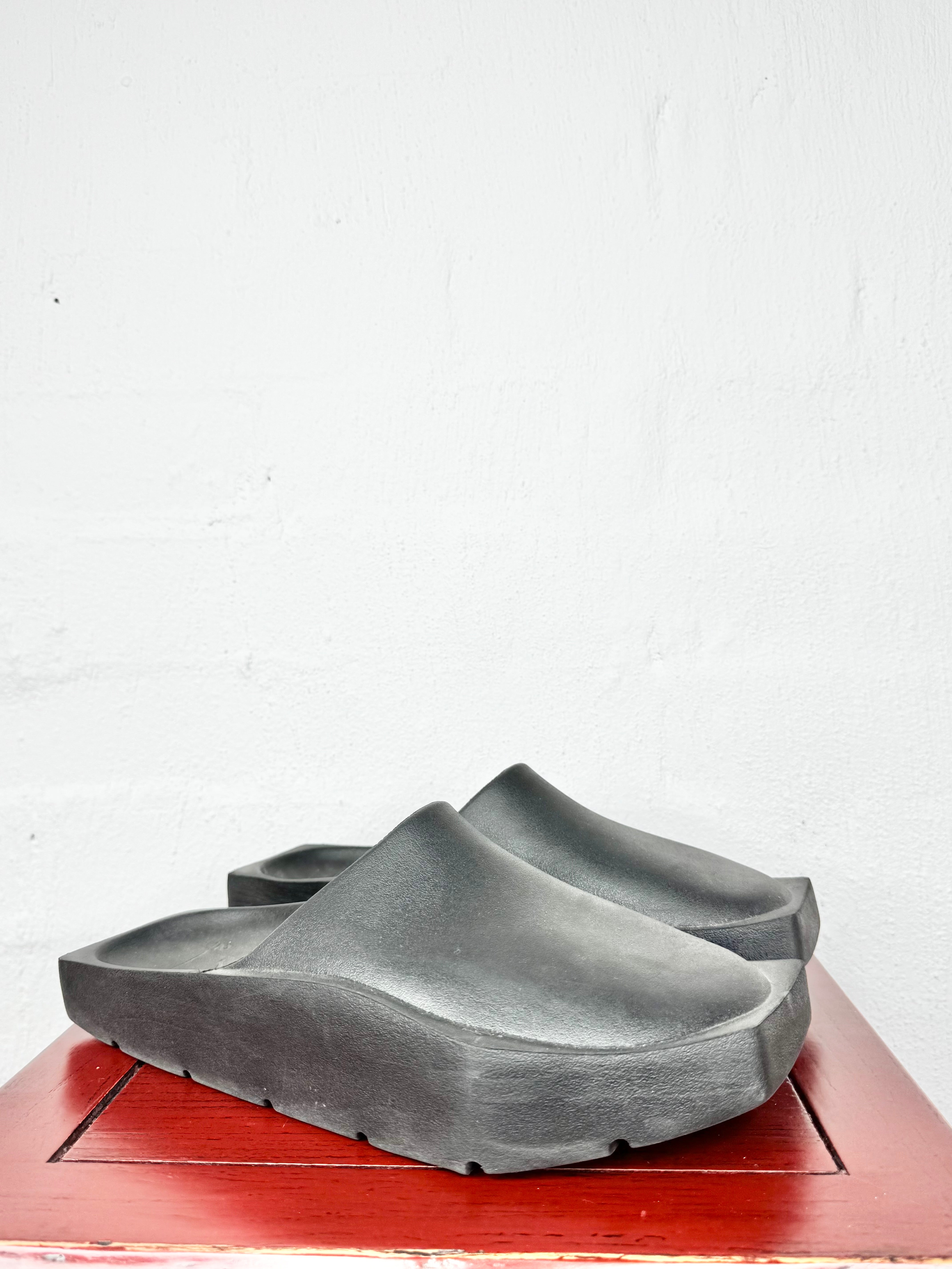 A pair of dark gray molded clogs with thick sculptural soles, displayed on a reddish-brown wooden surface against a white textured wall.
