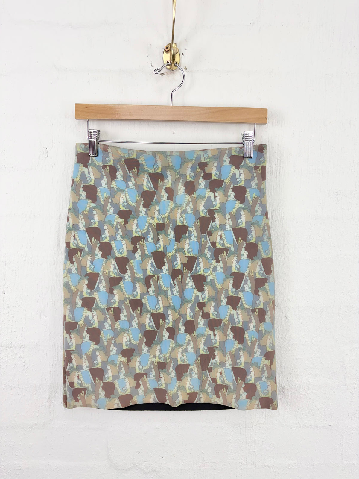 A patterned mini skirt with an abstract geometric print in brown, beige, and light blue, hanging from a wooden hanger against a white brick wall.