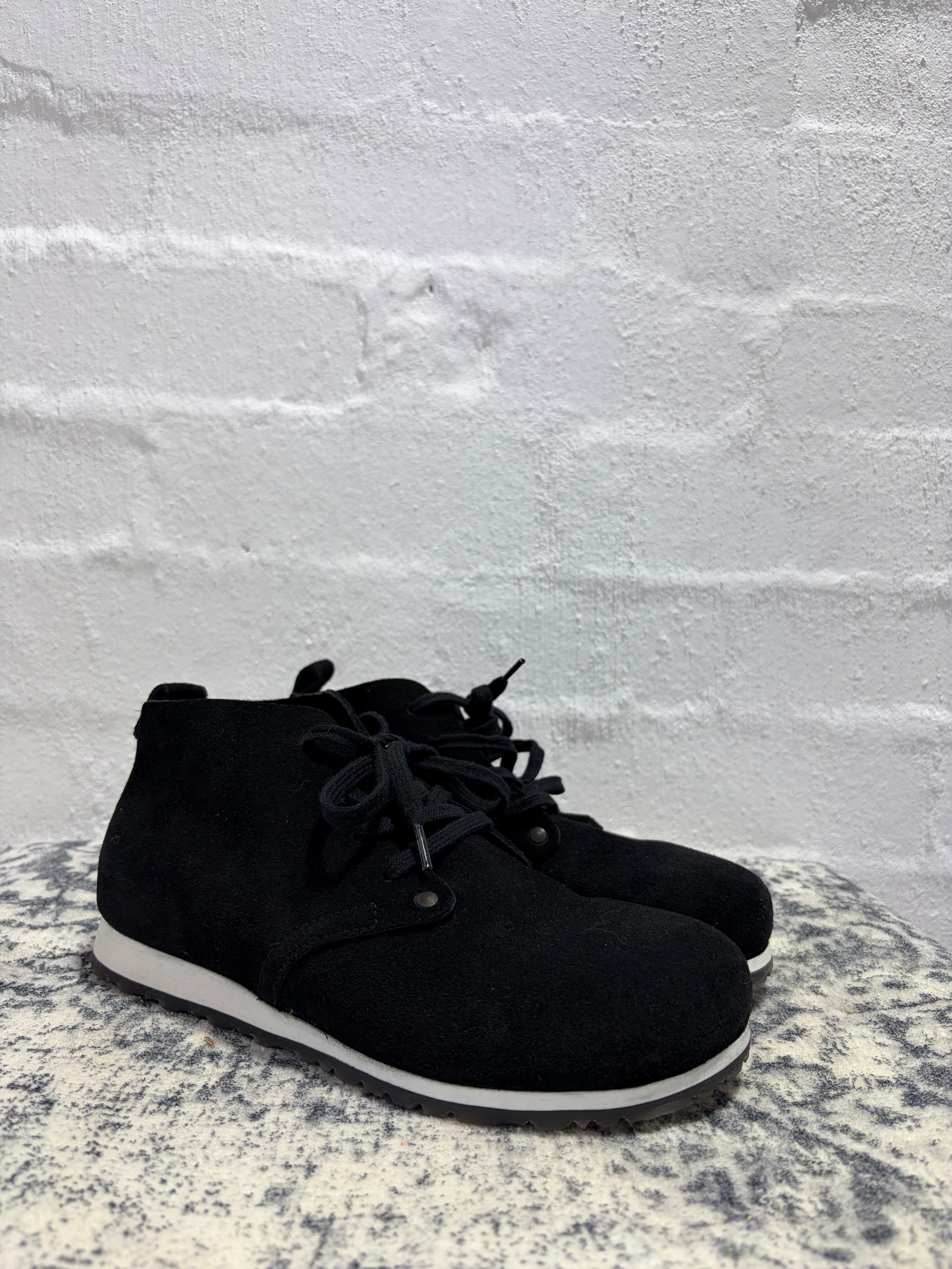 A pair of black suede ankle boots with black laces and white soles, photographed on a textured white brick wall.