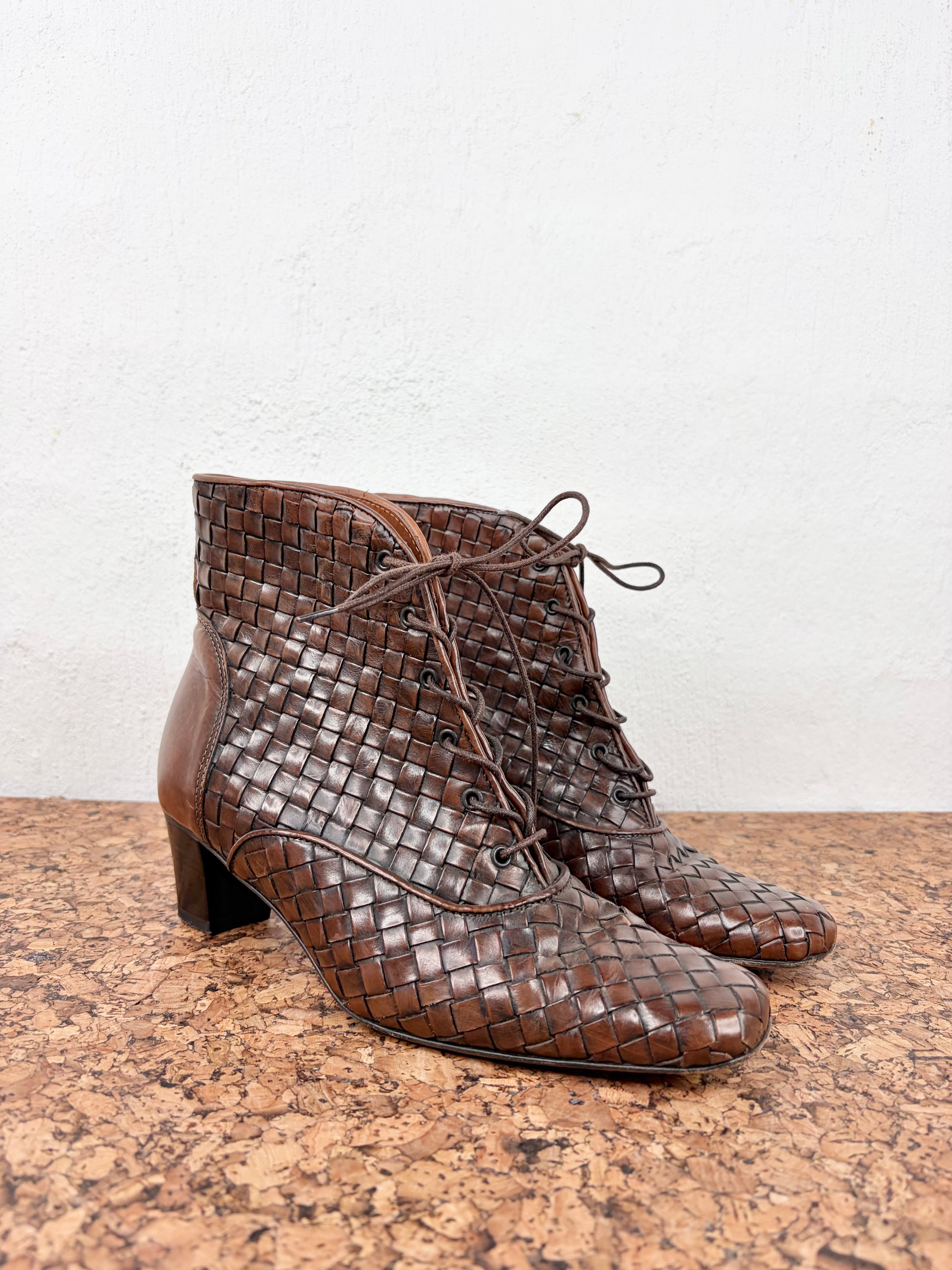 A pair of brown woven leather lace-up ankle boots with a low block heel, displayed on a cork surface against a white wall.