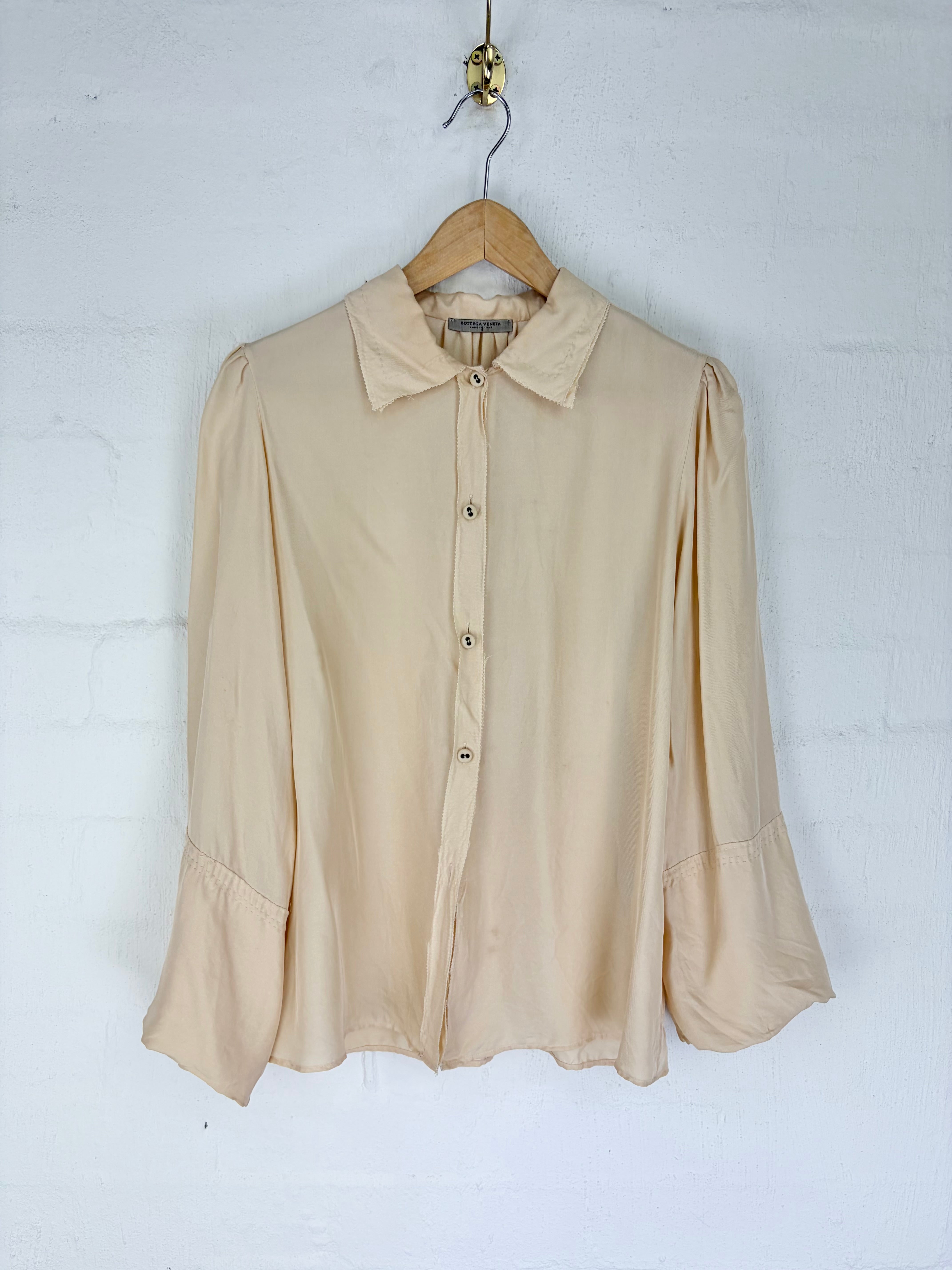 A light beige long-sleeve blouse with a pointed collar and button front, hanging on a wooden hanger against a white painted brick wall.