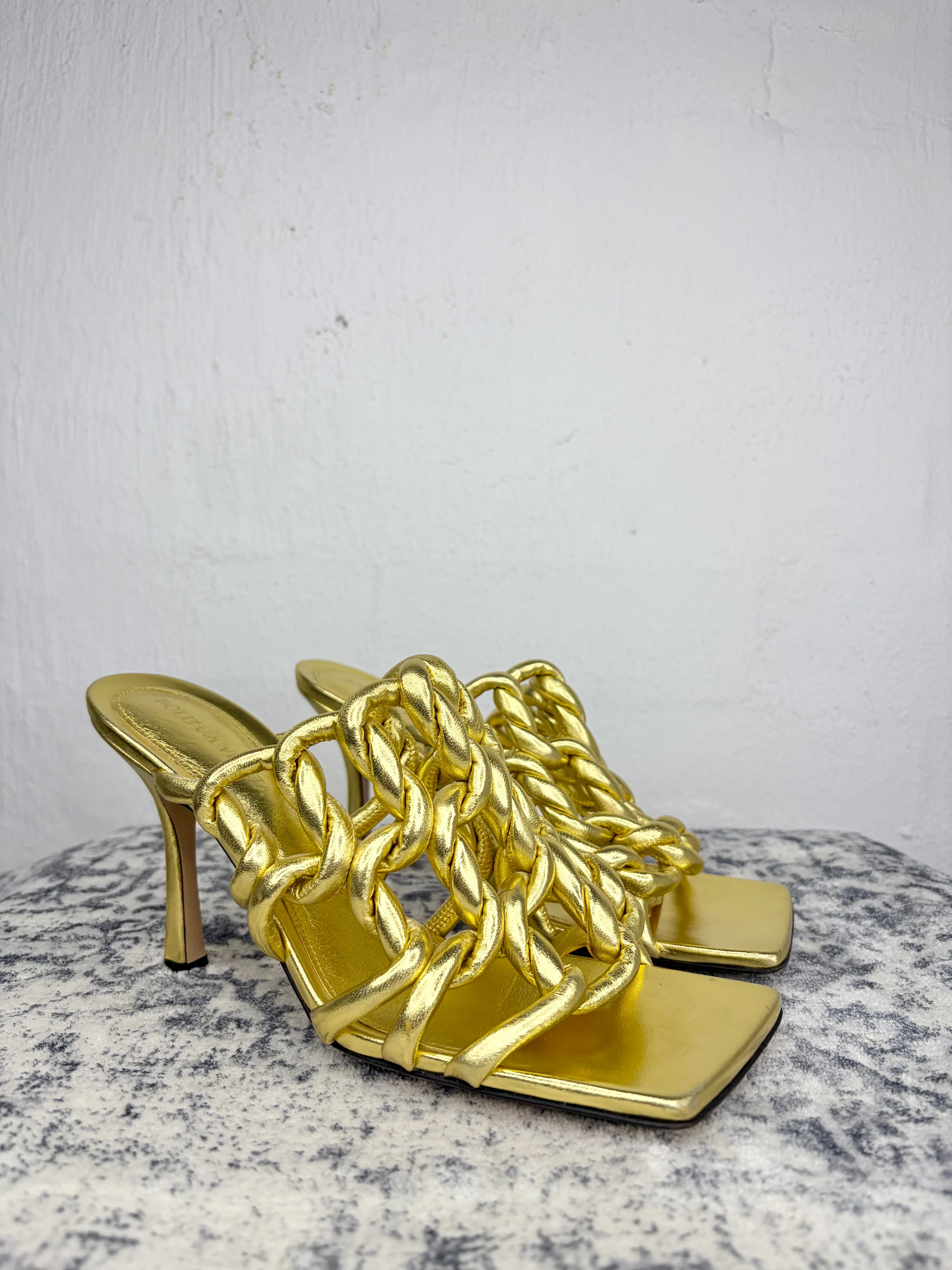 A pair of metallic gold high-heeled mule sandals with an intricate braided strap design, displayed on a speckled gray stone surface against a white wall.
