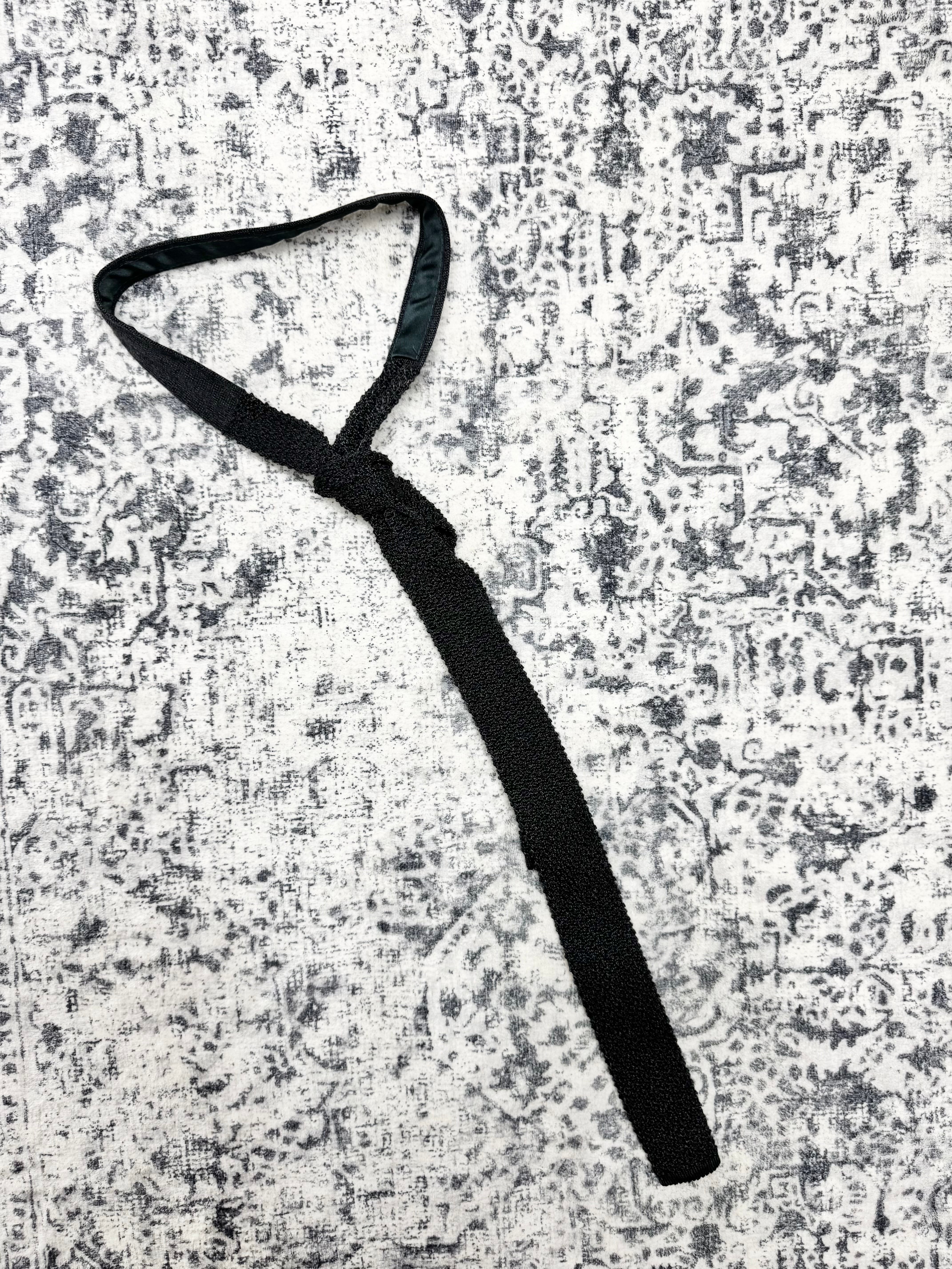 A black fabric strap accessory with a knotted loop, laid flat on a distressed gray and white patterned background.