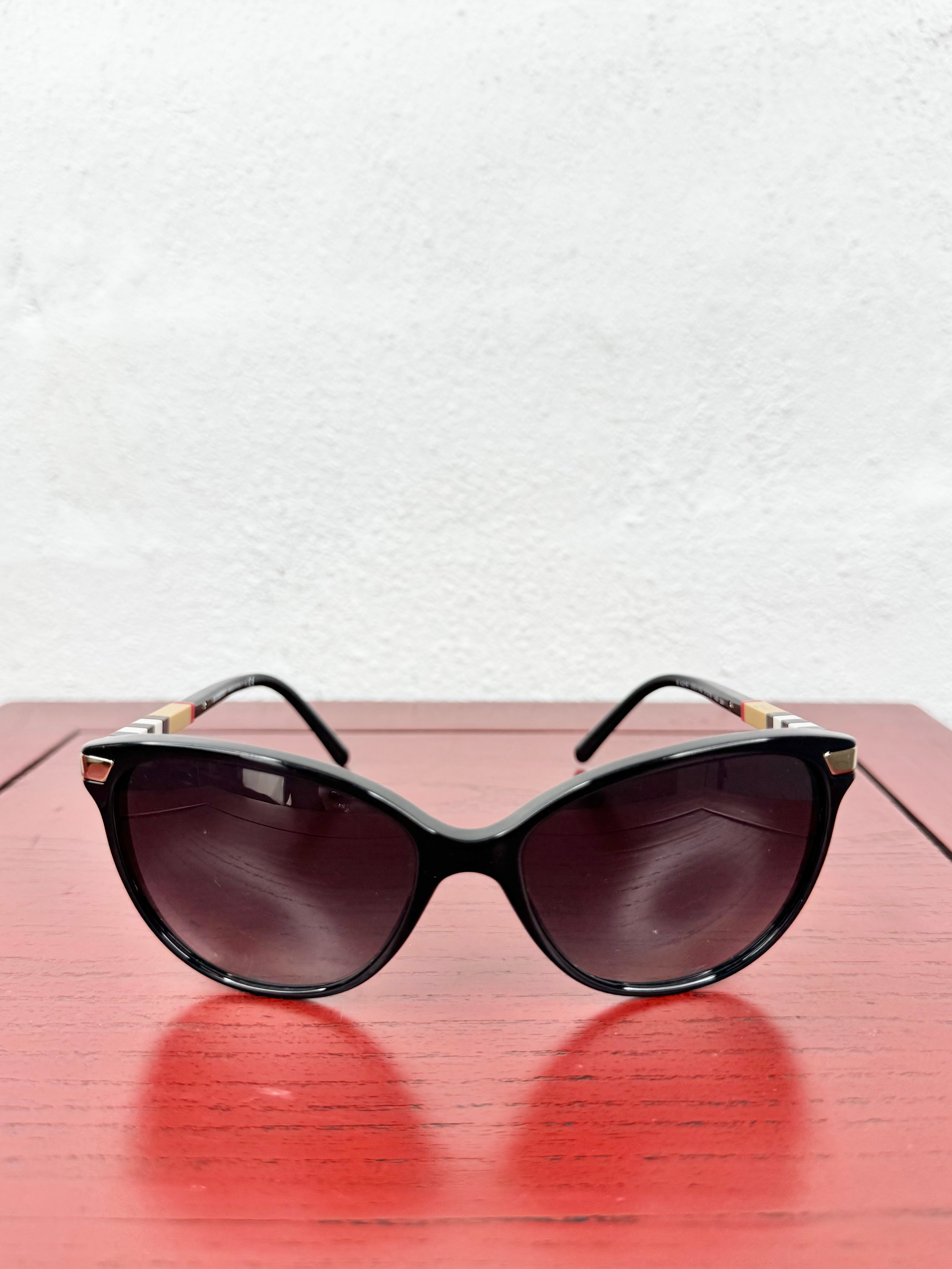 A pair of black cat-eye sunglasses with dark tinted lenses and small gold metal accents on the temples, resting on a red wooden surface against a white textured wall.