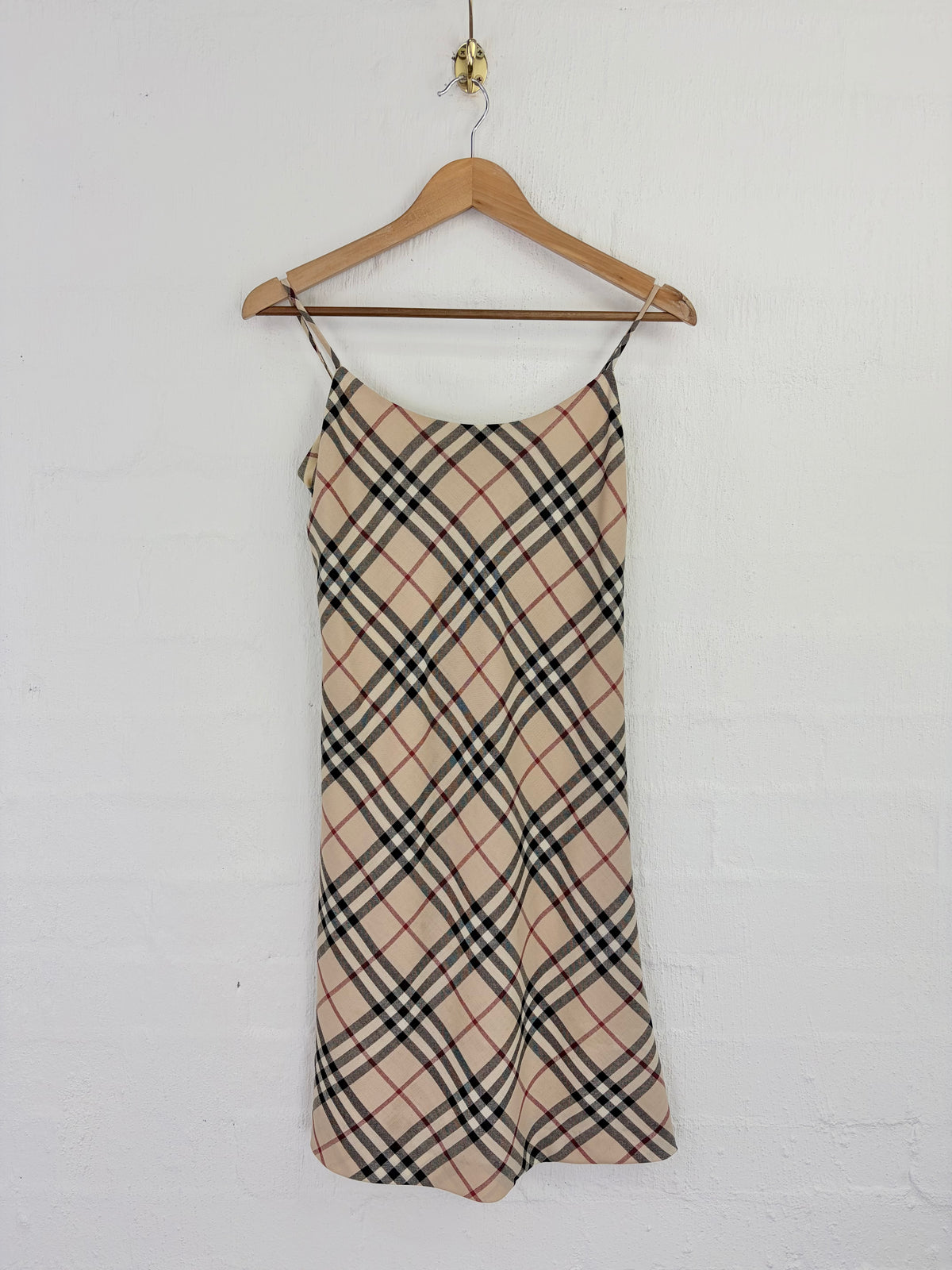 A beige plaid slip dress with thin straps, hanging on a wooden hanger against a white painted brick wall.