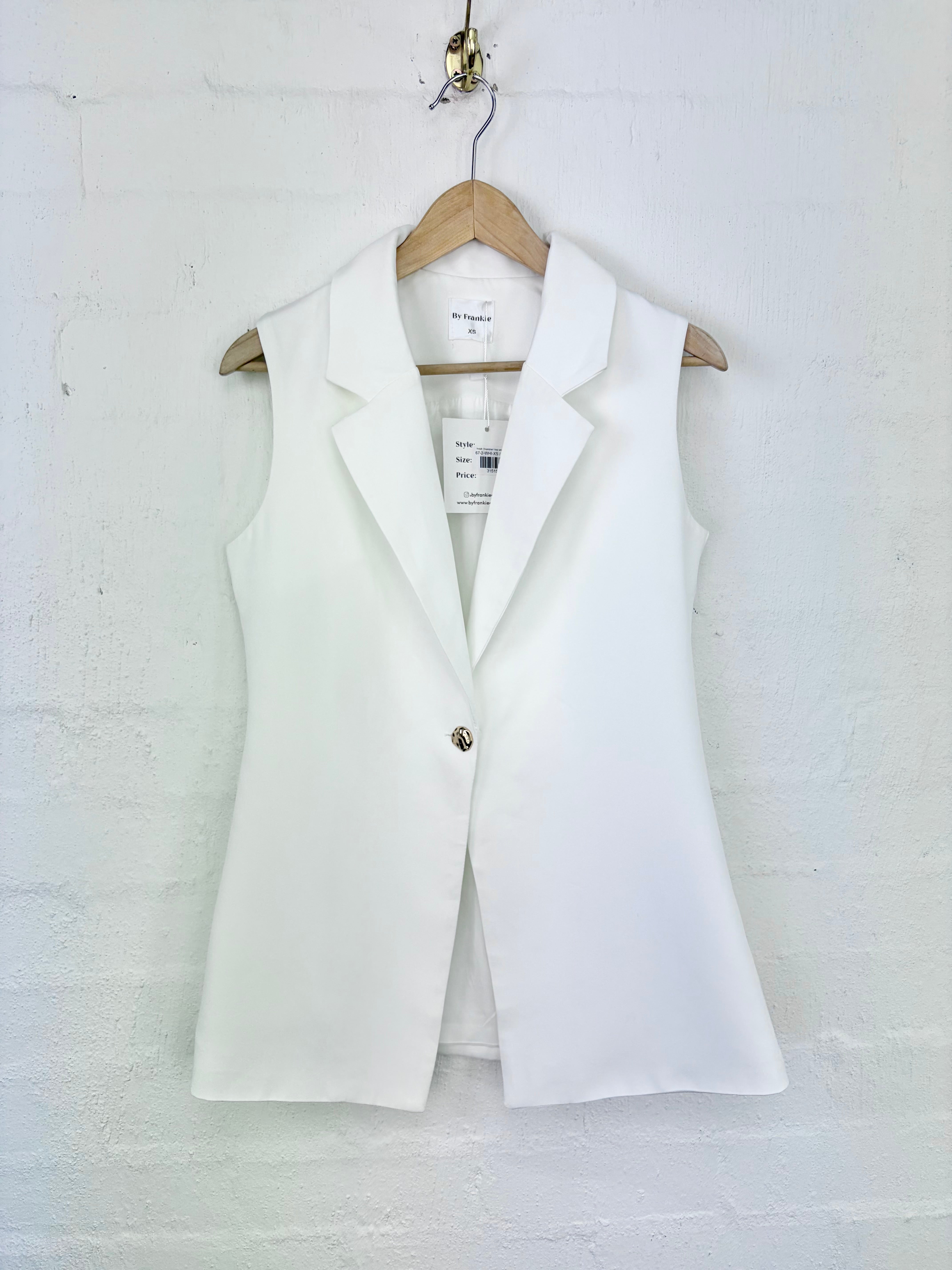 A white sleeveless blazer vest with a single gold button, hanging on a wooden hanger against a white painted brick wall.