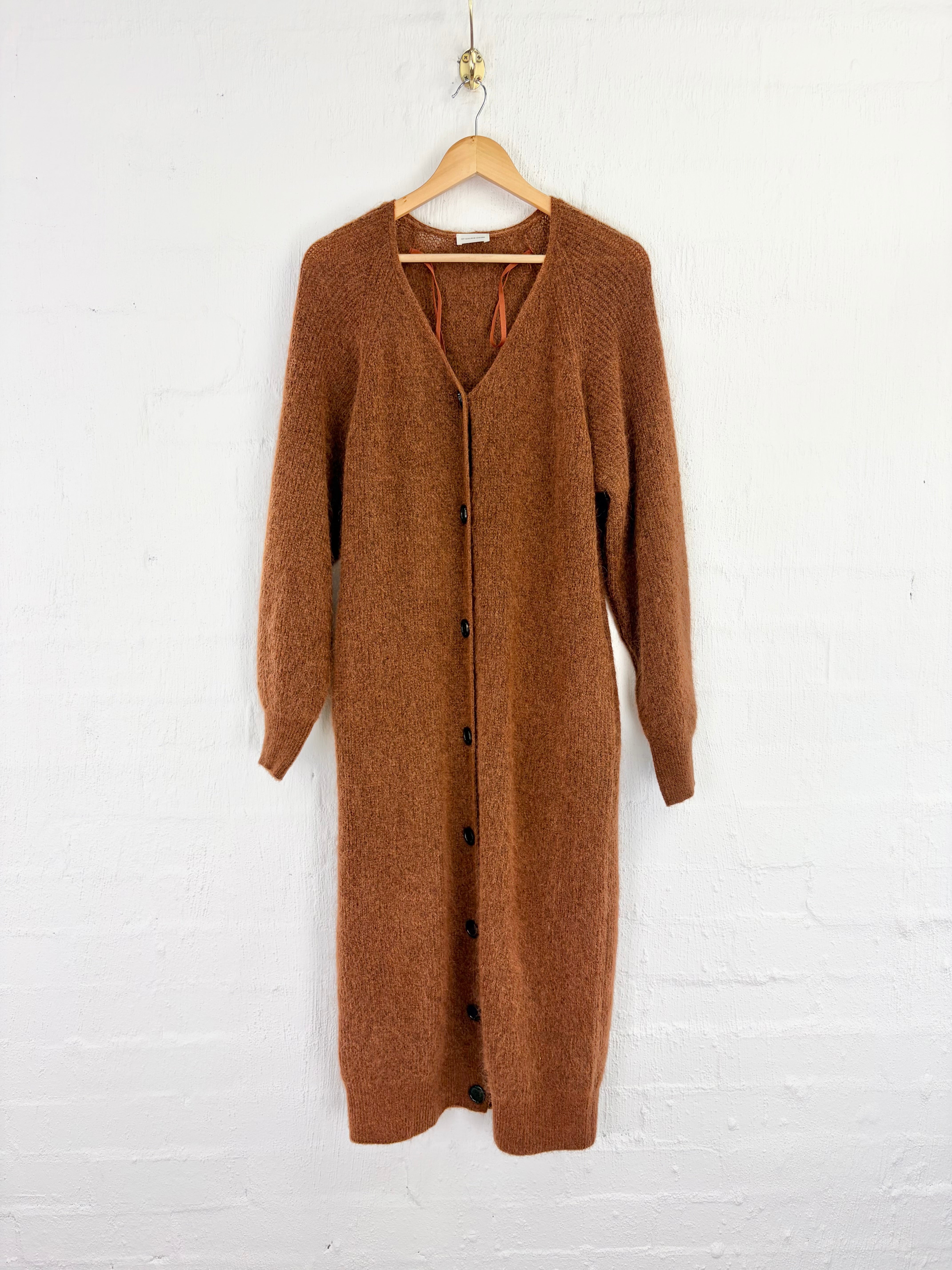 A brown knit cardigan dress with a V-neck and front buttons, hanging on a wooden hanger against a white brick wall.
