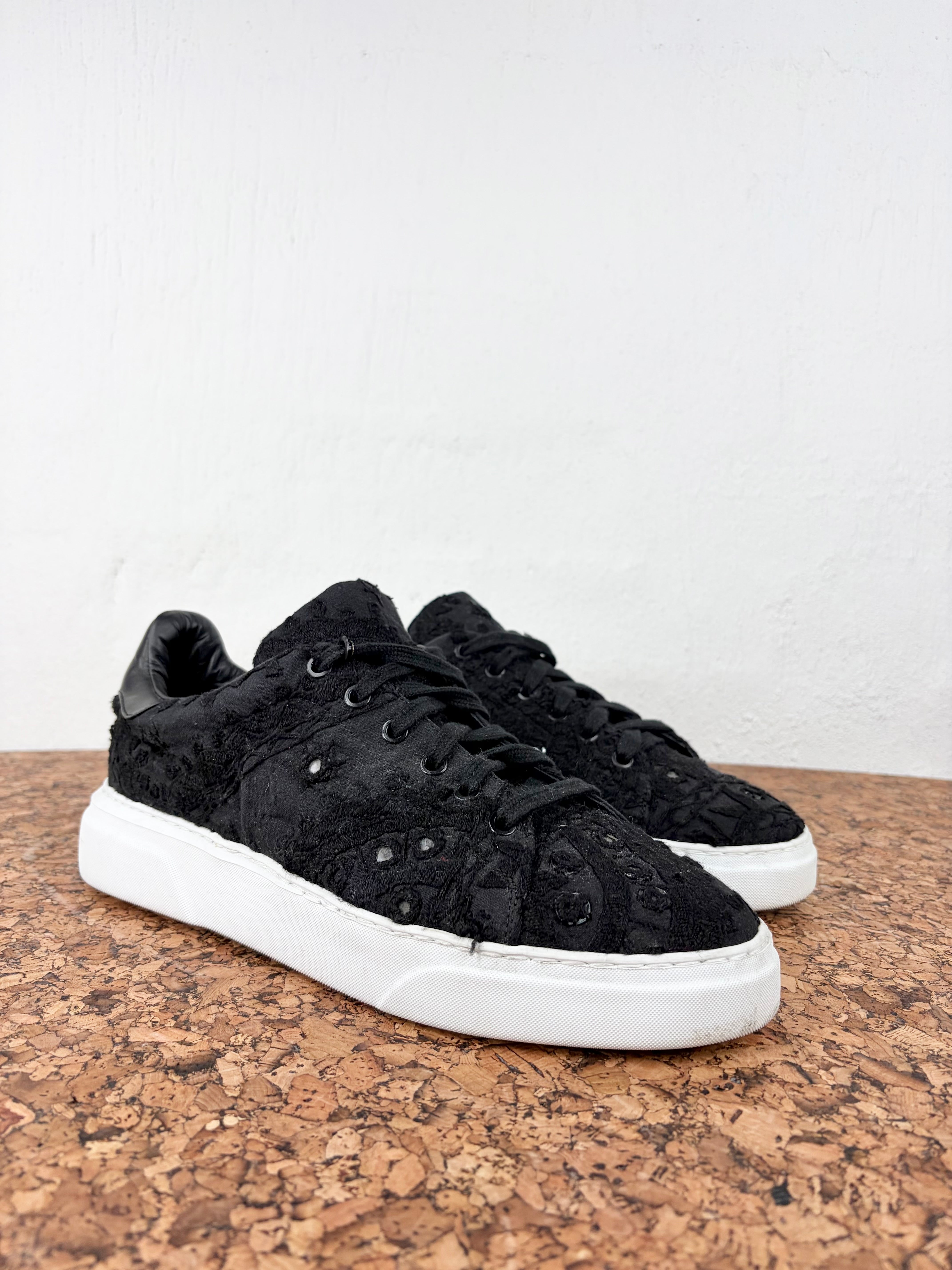 A pair of black textured lace-up sneakers with white platform soles, displayed on a cork surface against a white wall.