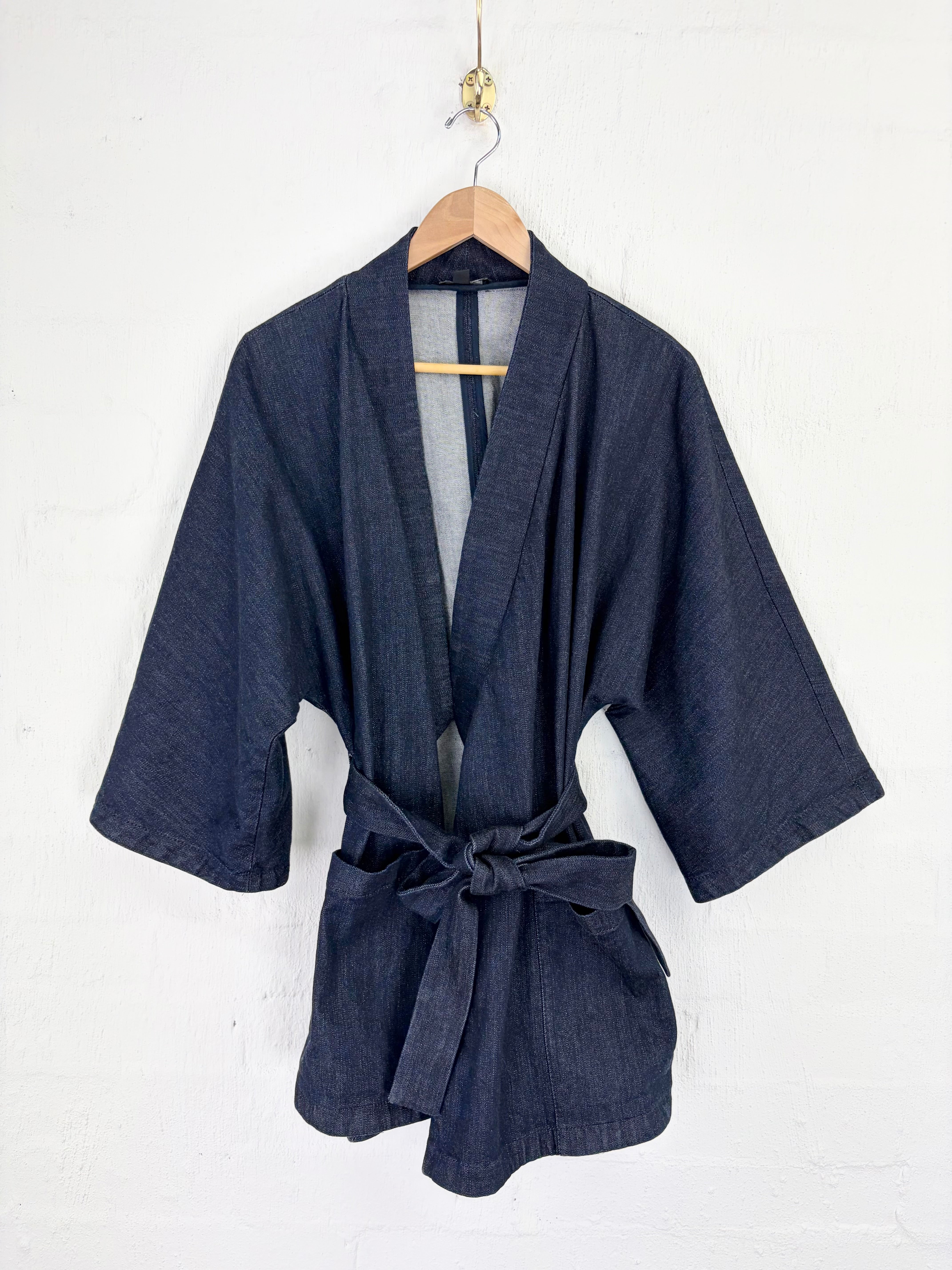 A dark navy blue kimono-style robe with three-quarter sleeves and a self-tie belt, hanging on a wooden hanger against a white painted brick wall.