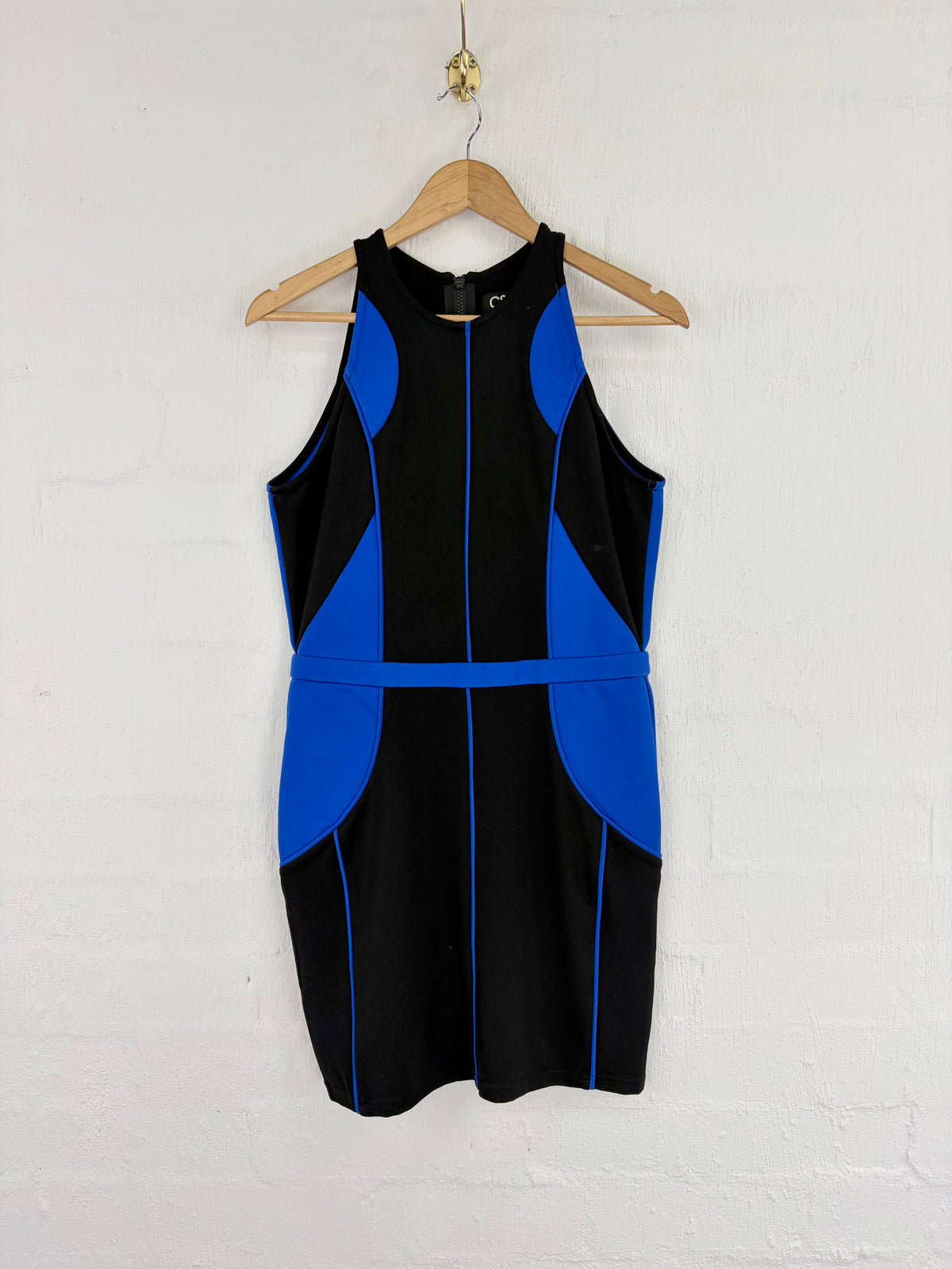 A sleeveless black and royal blue color-block dress hanging on a wooden hanger against a white painted brick wall.