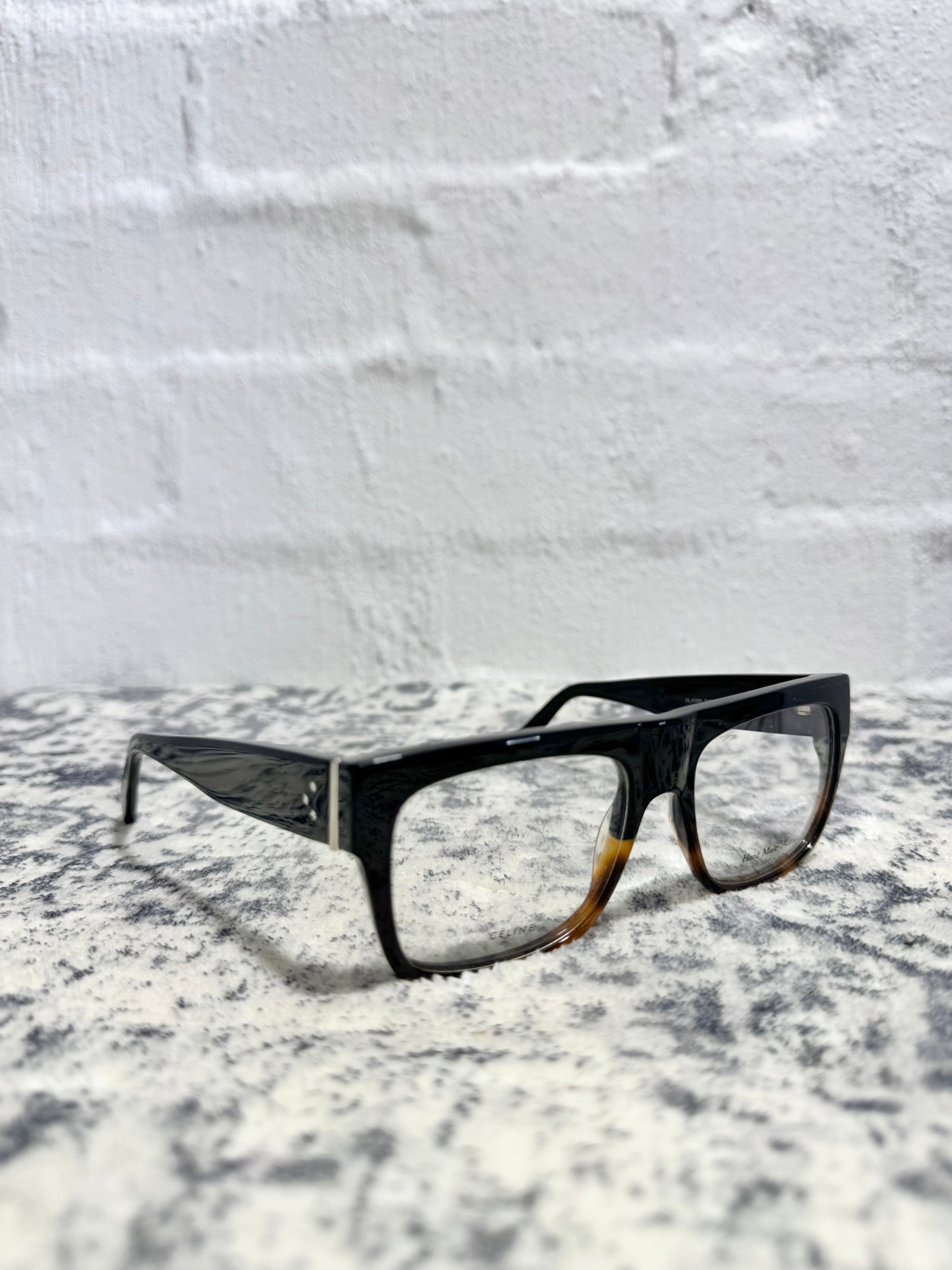 A pair of square-frame eyeglasses with black acetate fronts and tortoiseshell-patterned lower rims, resting on a textured gray and white surface against a white brick wall background.