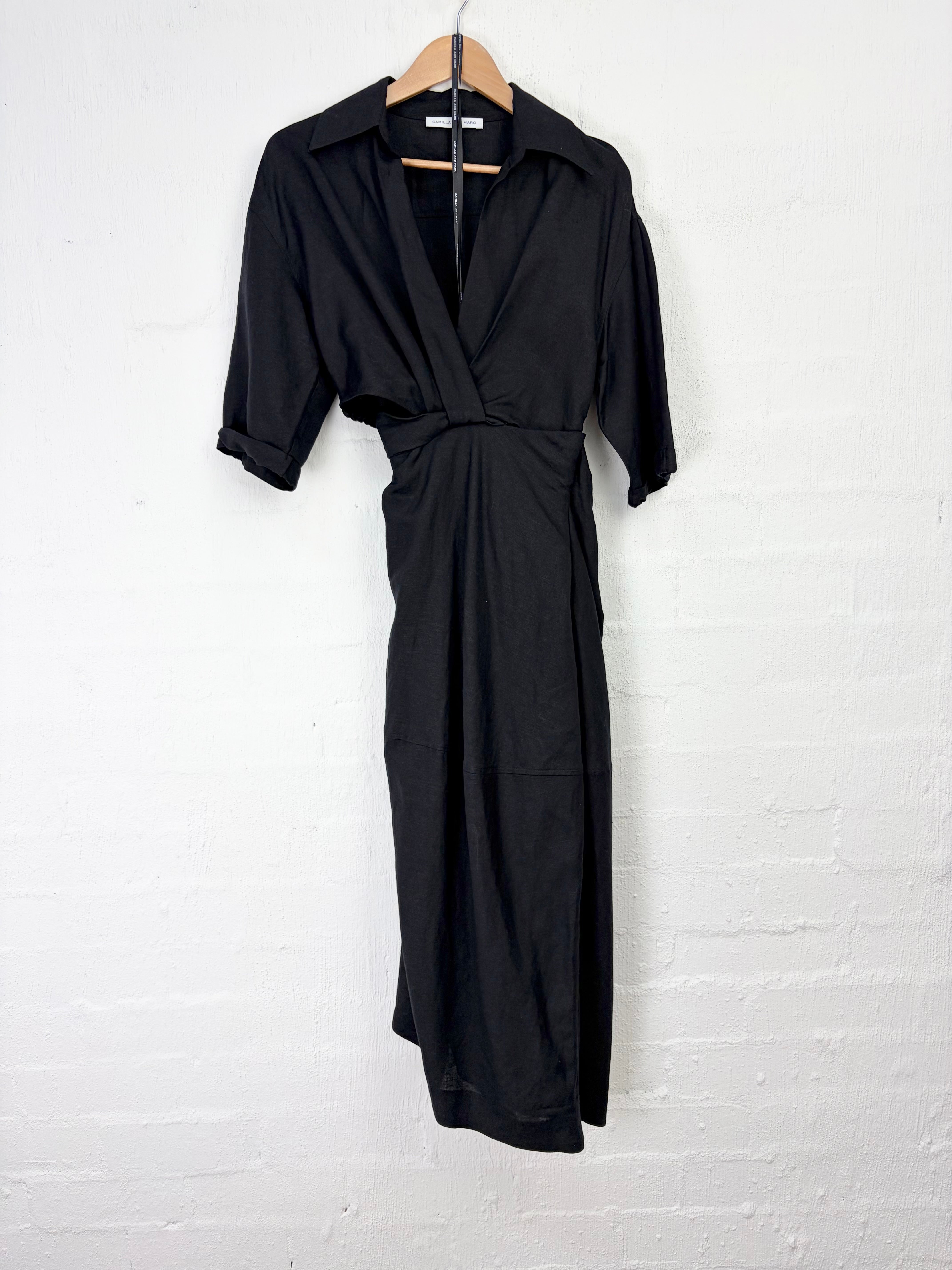 A black short-sleeve wrap-style midi dress hanging on a wooden hanger against a white painted brick wall.