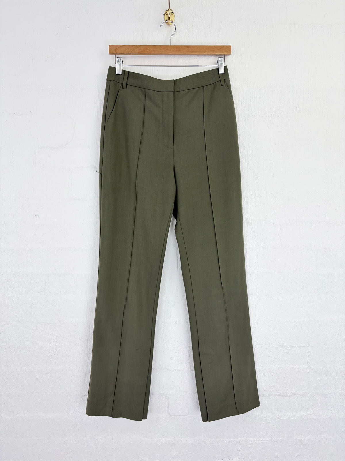 A pair of olive green wide-leg trousers hanging from a wooden hanger against a white painted brick wall.