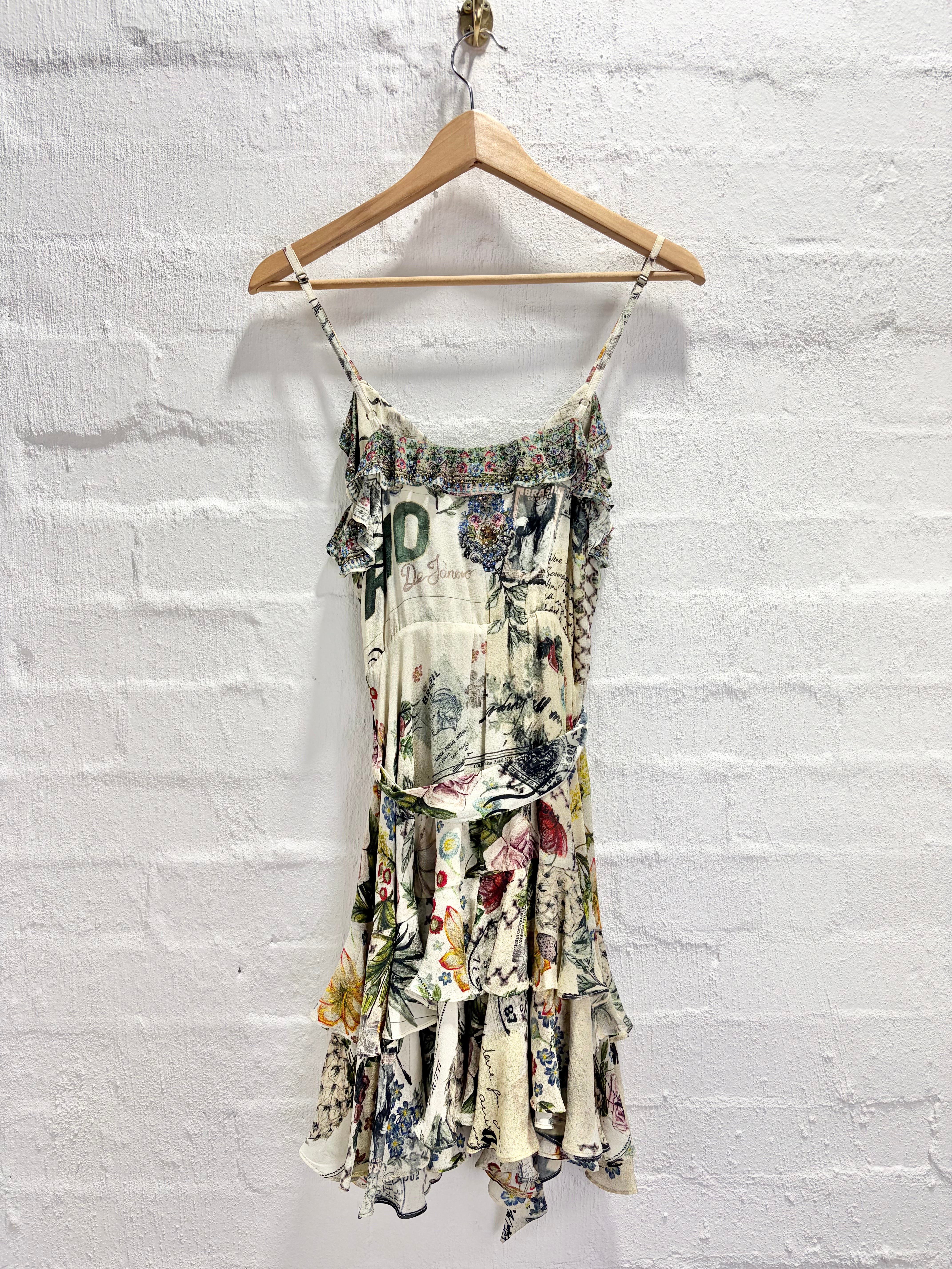 A sleeveless ruffled mini dress with a multicolor floral and botanical print on a cream base, hanging on a wooden hanger against a white painted brick wall.