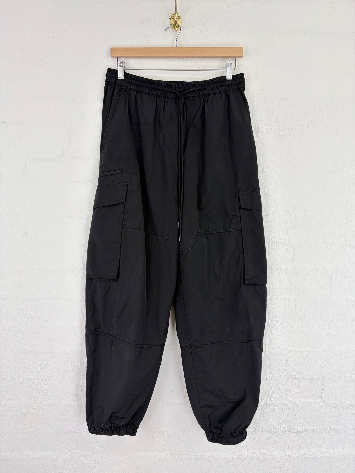 Black cargo jogger pants with side flap pockets and elastic cuffs, hanging from a wooden hanger against a white painted brick wall.