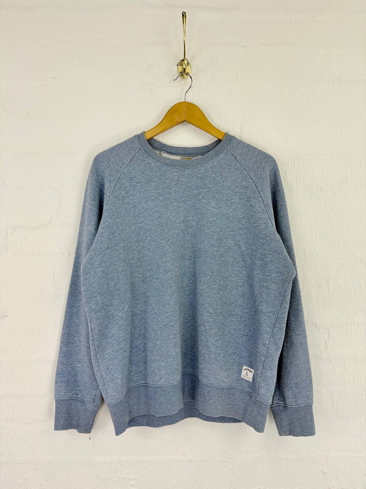 A heathered blue crewneck sweatshirt hanging on a wooden hanger against a white textured wall.