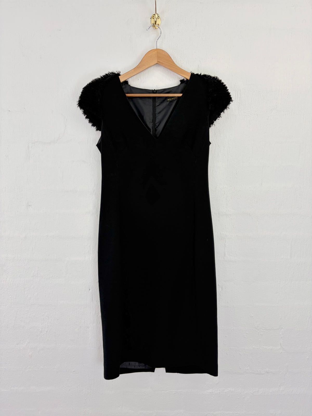 A black sleeveless dress with fuzzy cap sleeves and a deep V-neckline, hanging on a wooden hanger against a white painted brick wall.
