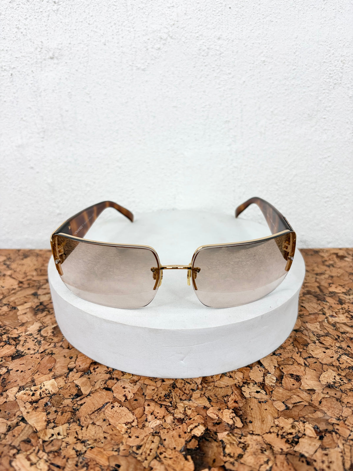 A pair of rimless square sunglasses with gold metal frames and brown gradient lenses, resting on a white cylindrical display stand over a cork surface against a white textured wall.