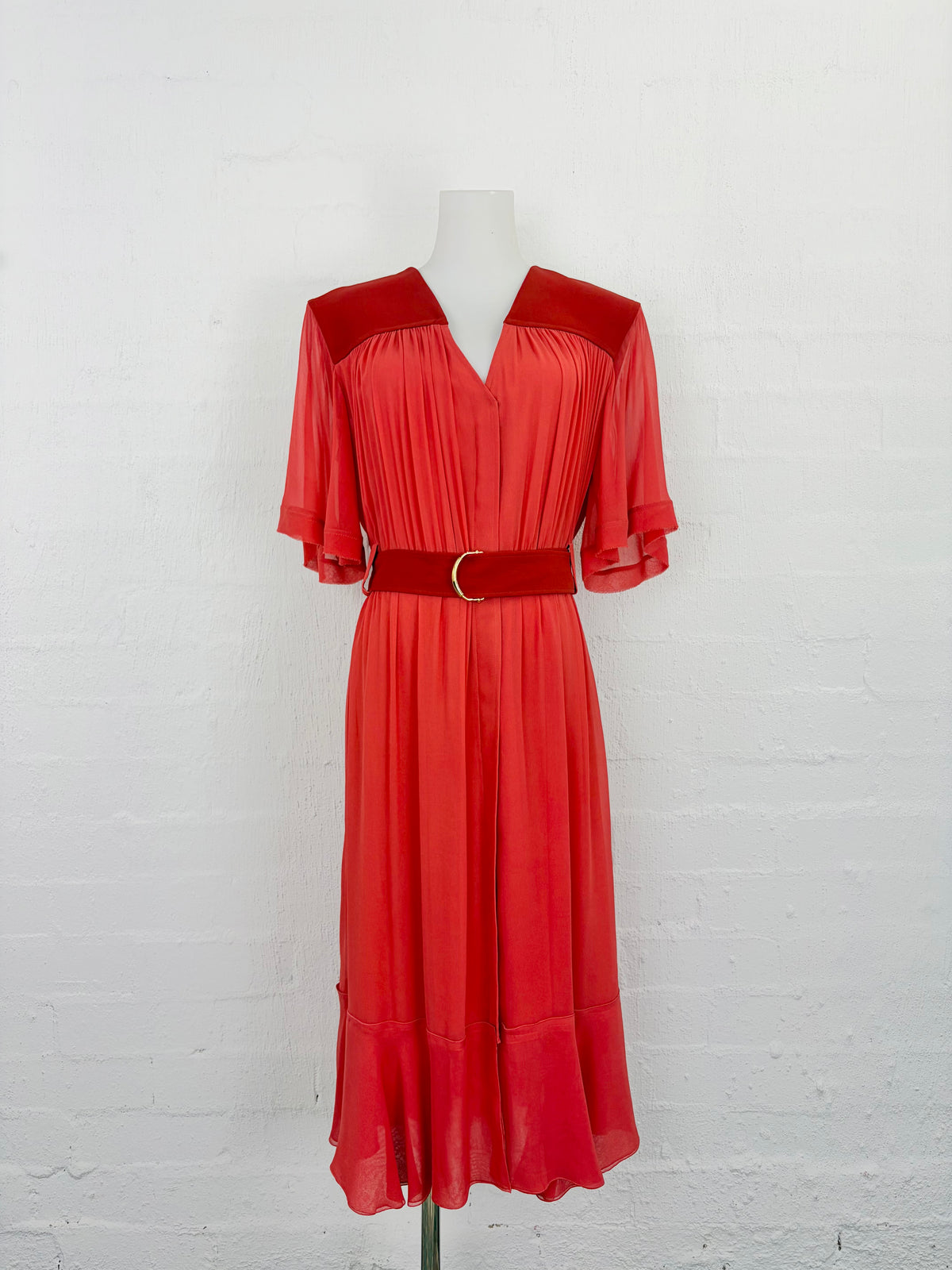 A red midi dress with a structured velvet-like collar and shoulder panels, short flutter sleeves, a matching waist belt with a gold-tone ring, and a pleated skirt, displayed on a mannequin against a white brick wall.