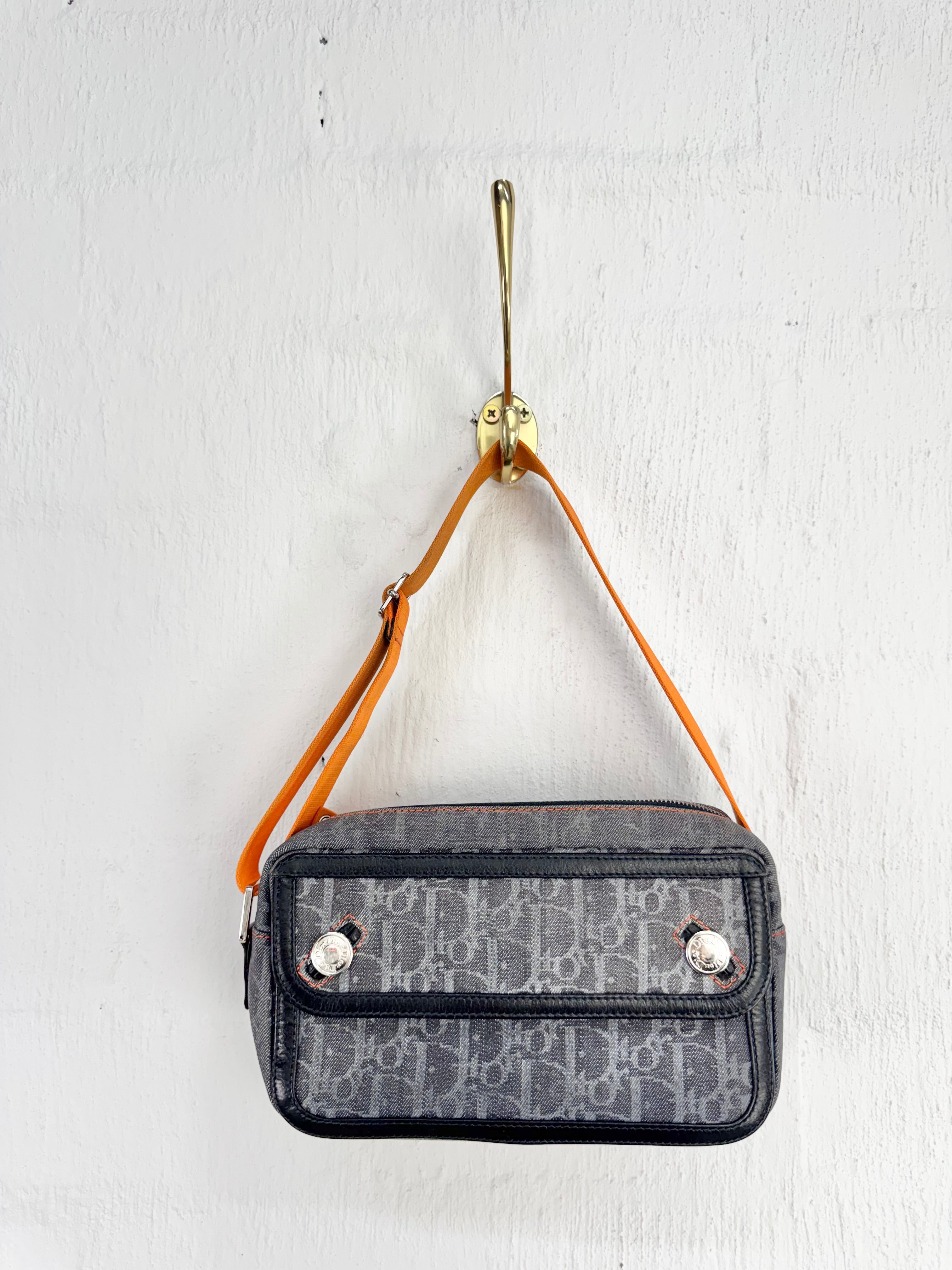 A small Dior monogram shoulder bag in gray and black, hanging from a gold hook on a white textured wall. The bag features a signature Dior Oblique monogram pattern, black leather trim, and a tan leather shoulder strap.