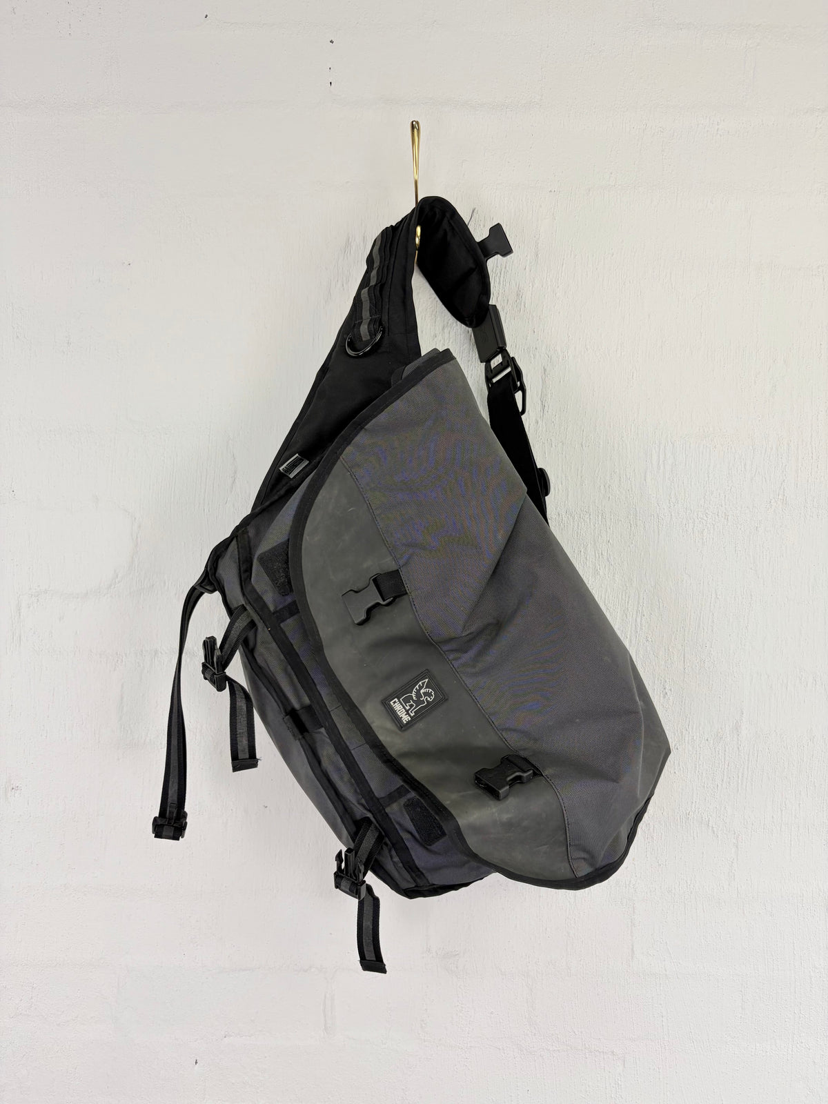 A gray and black sling backpack with multiple straps and buckles, hanging from a small hook on a white painted brick wall.