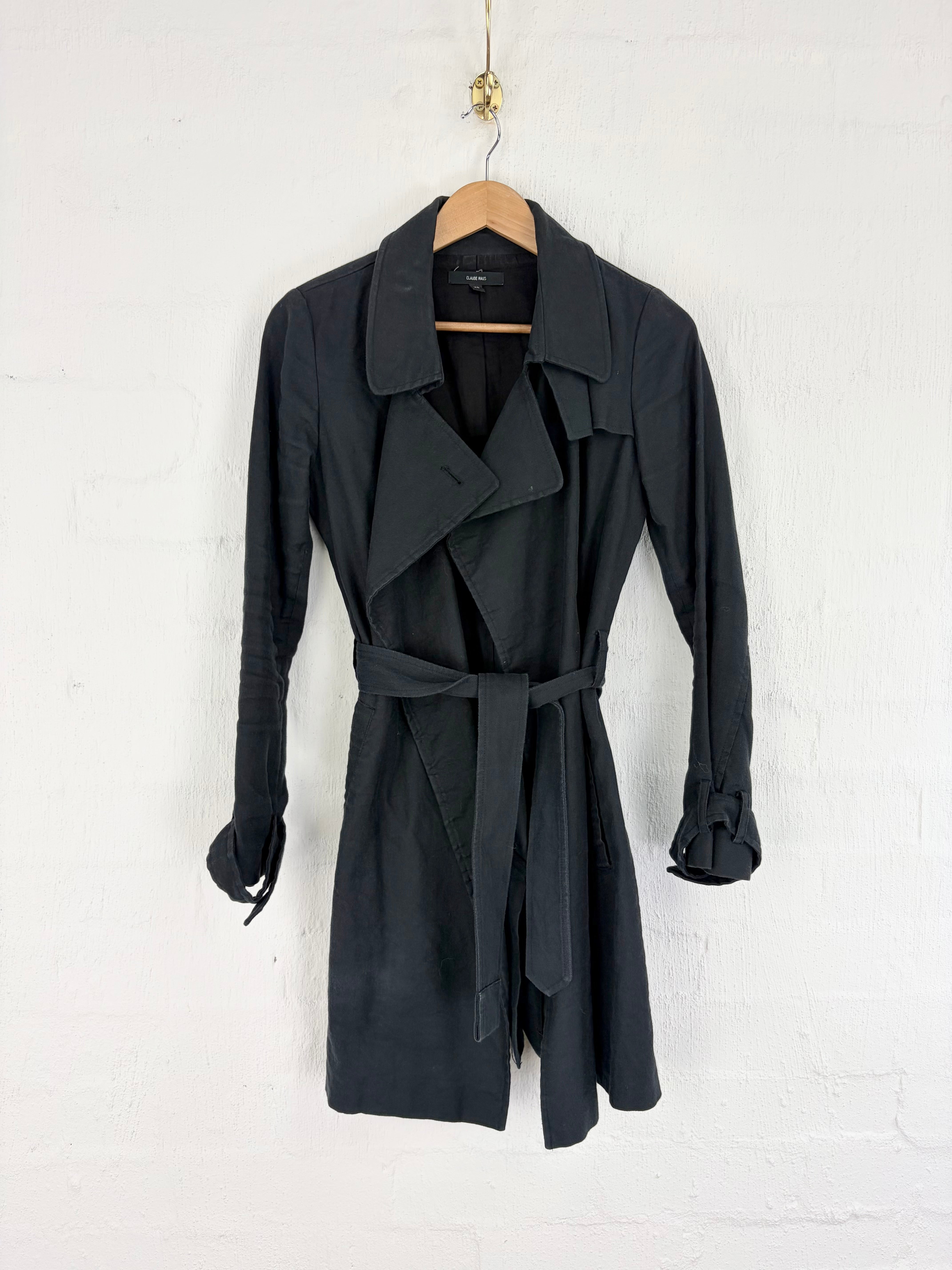 A black trench coat with a matching waist tie belt, hanging on a wooden hanger against a white painted brick wall.
