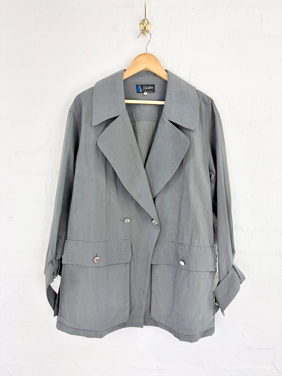 A gray double-breasted blazer jacket with wide lapels and flap pockets, hanging on a wooden hanger against a white textured wall.