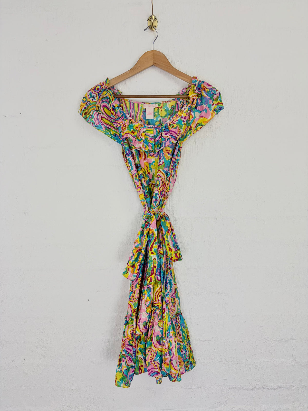 A colorful abstract print mini dress with puff sleeves and a tie waist, hanging on a wooden hanger against a white wall.