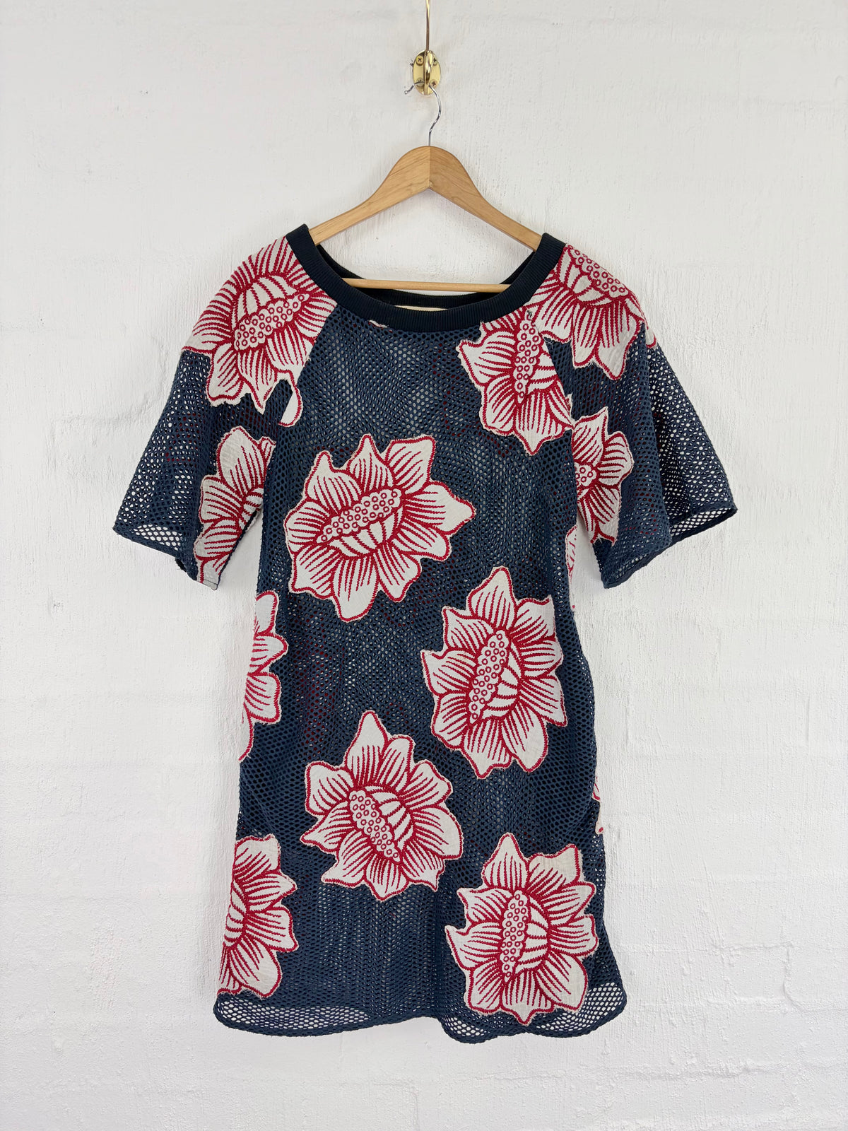 A short-sleeve mesh tunic dress with a navy blue base and large red and white floral print, hanging on a wooden hanger against a white brick wall.