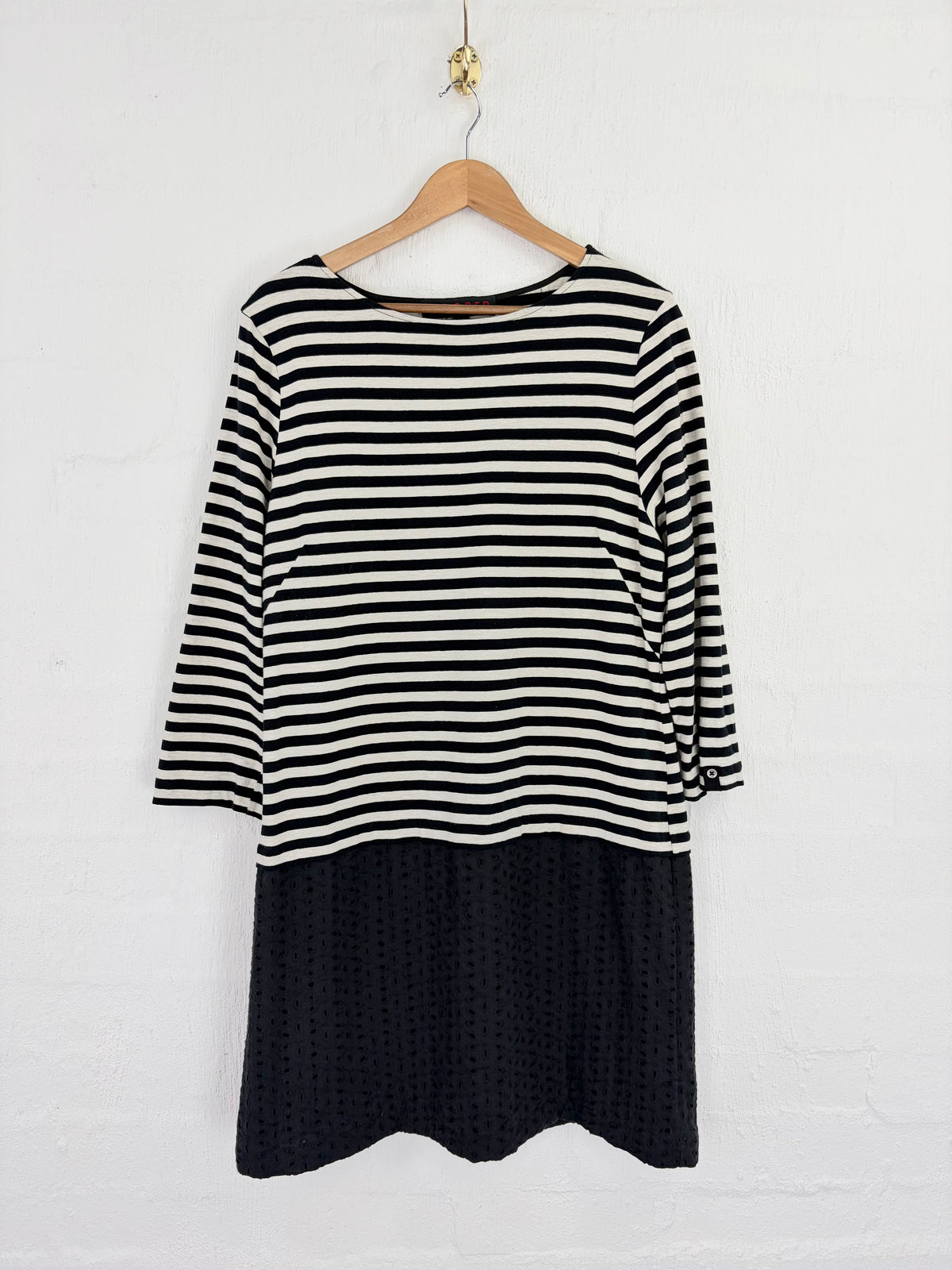 A women's long-sleeve dress hanging on a wooden hanger against a white brick wall. The top portion features horizontal black and off-white stripes, while the bottom portion is a solid black textured knit.