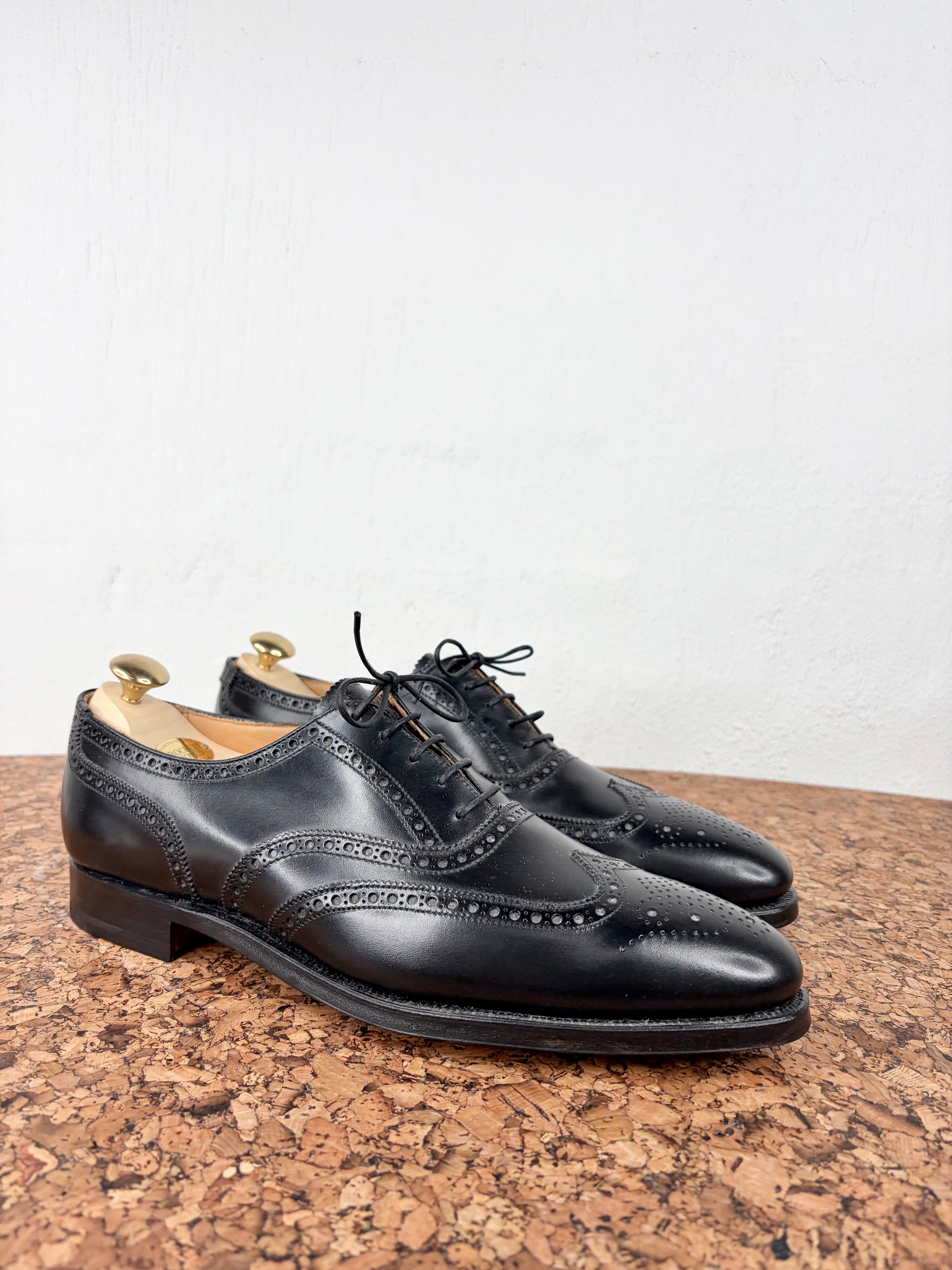 A pair of black leather wingtip oxford shoes with brogue detailing, displayed on cork shoe trees against a white wall.