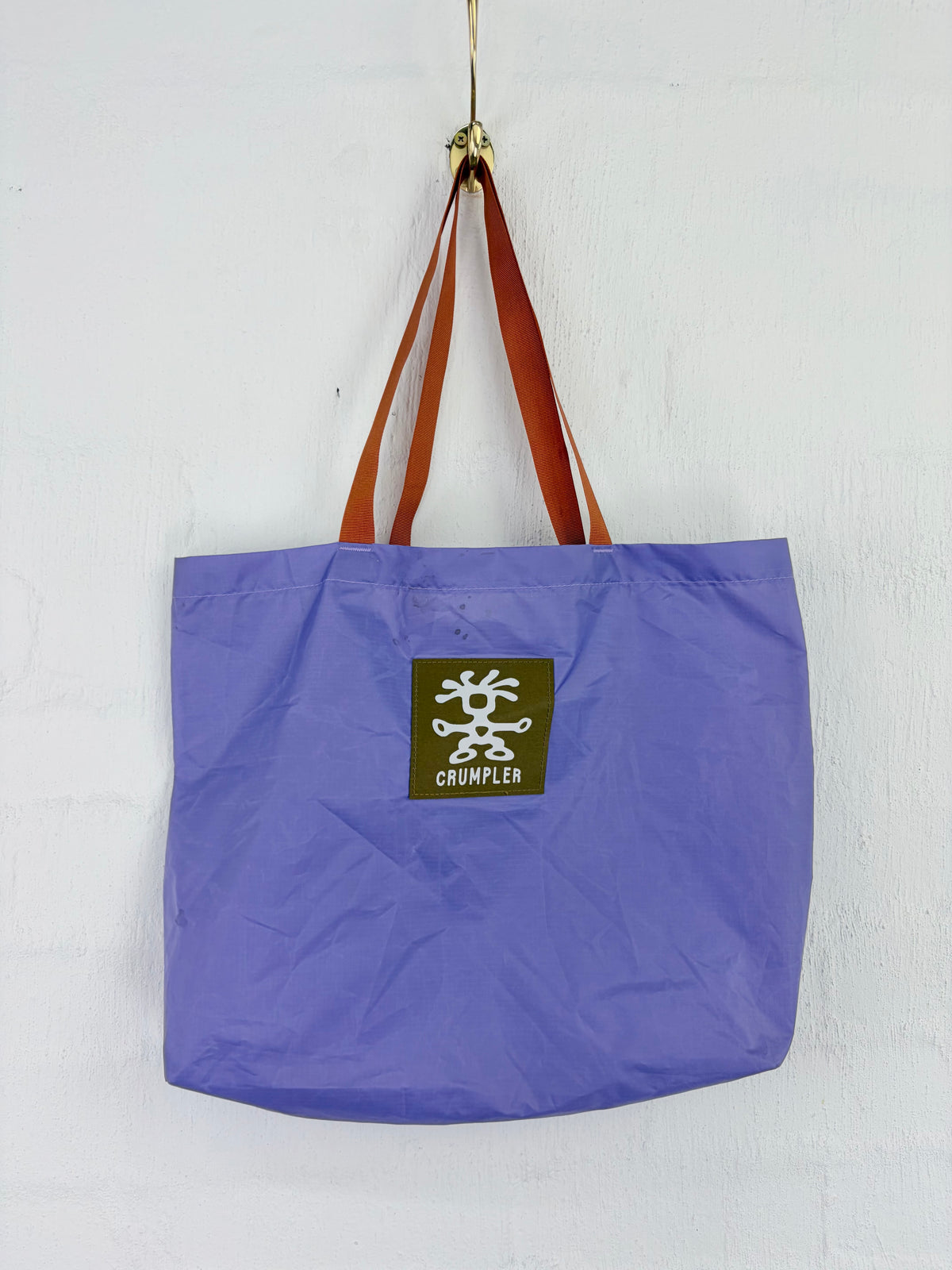A purple fabric tote bag with brown shoulder straps hanging from a gold hook against a white wall. A square brown patch on the front features a white stylized logo and the brand name 'CRUMBLE'.