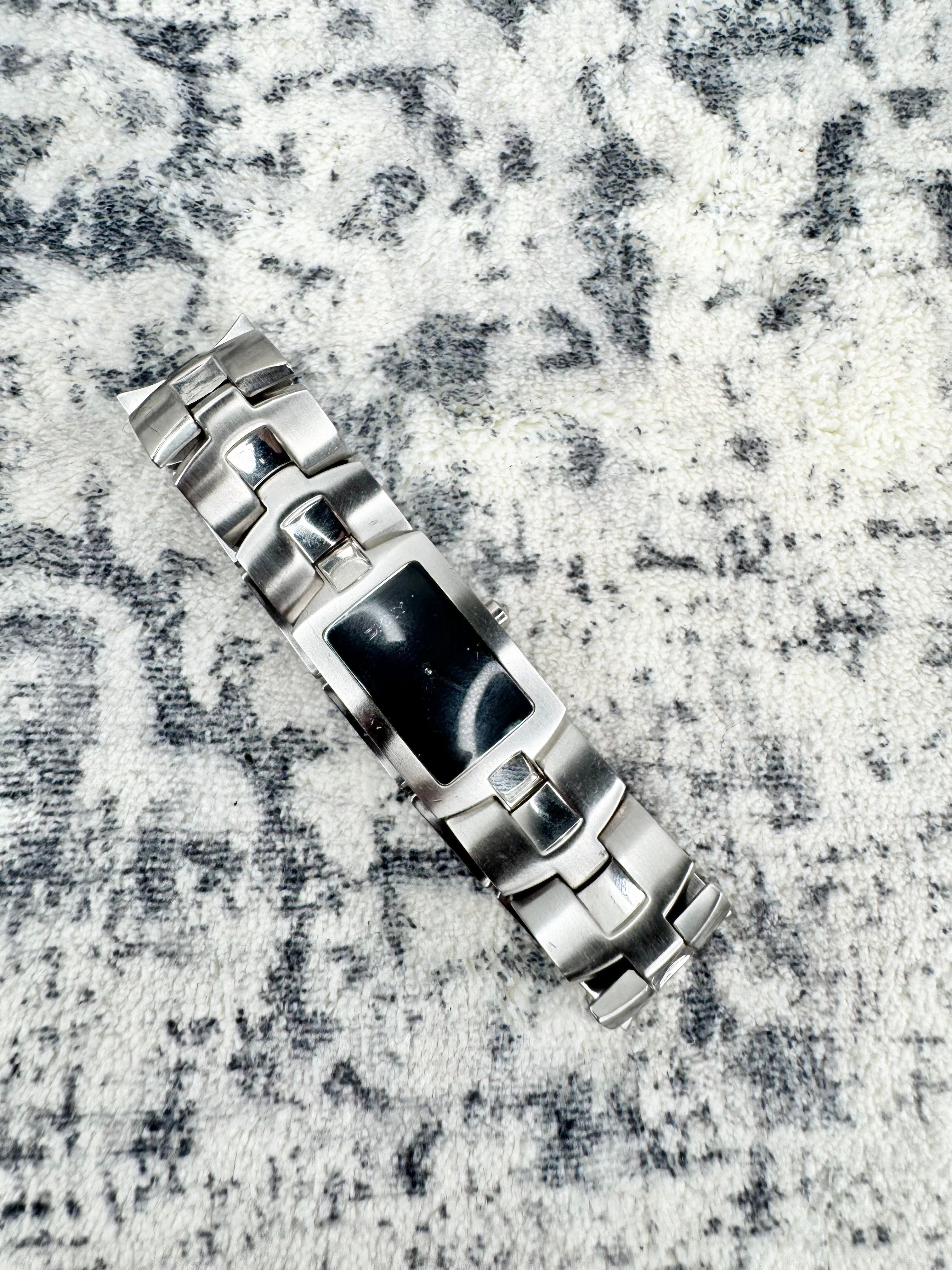 A silver stainless steel watch band with a rectangular black inlay, shown diagonally on a textured gray and white marbled surface.