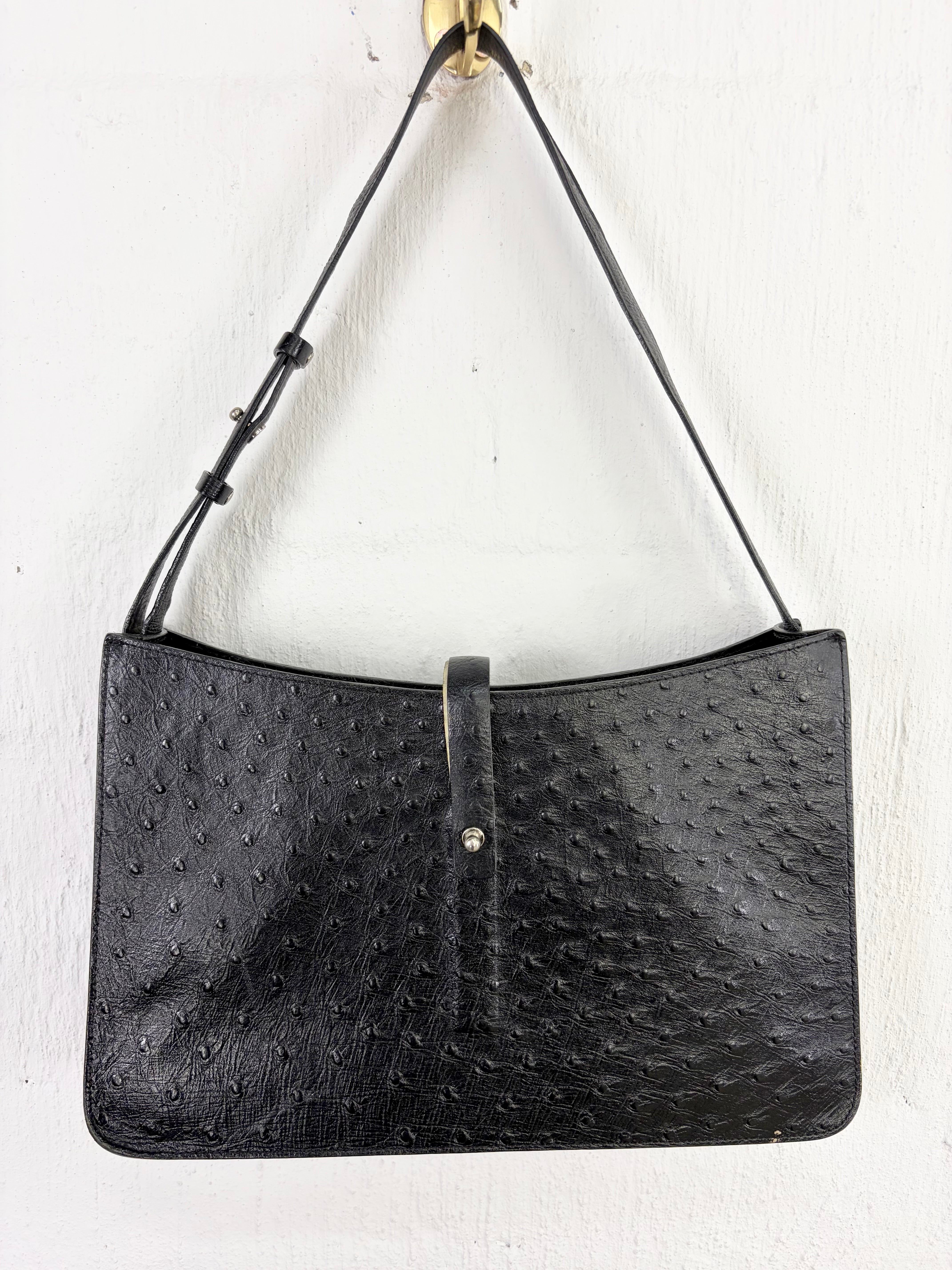 A black ostrich-embossed leather shoulder bag hanging from a gold wall hook against a white textured wall.
