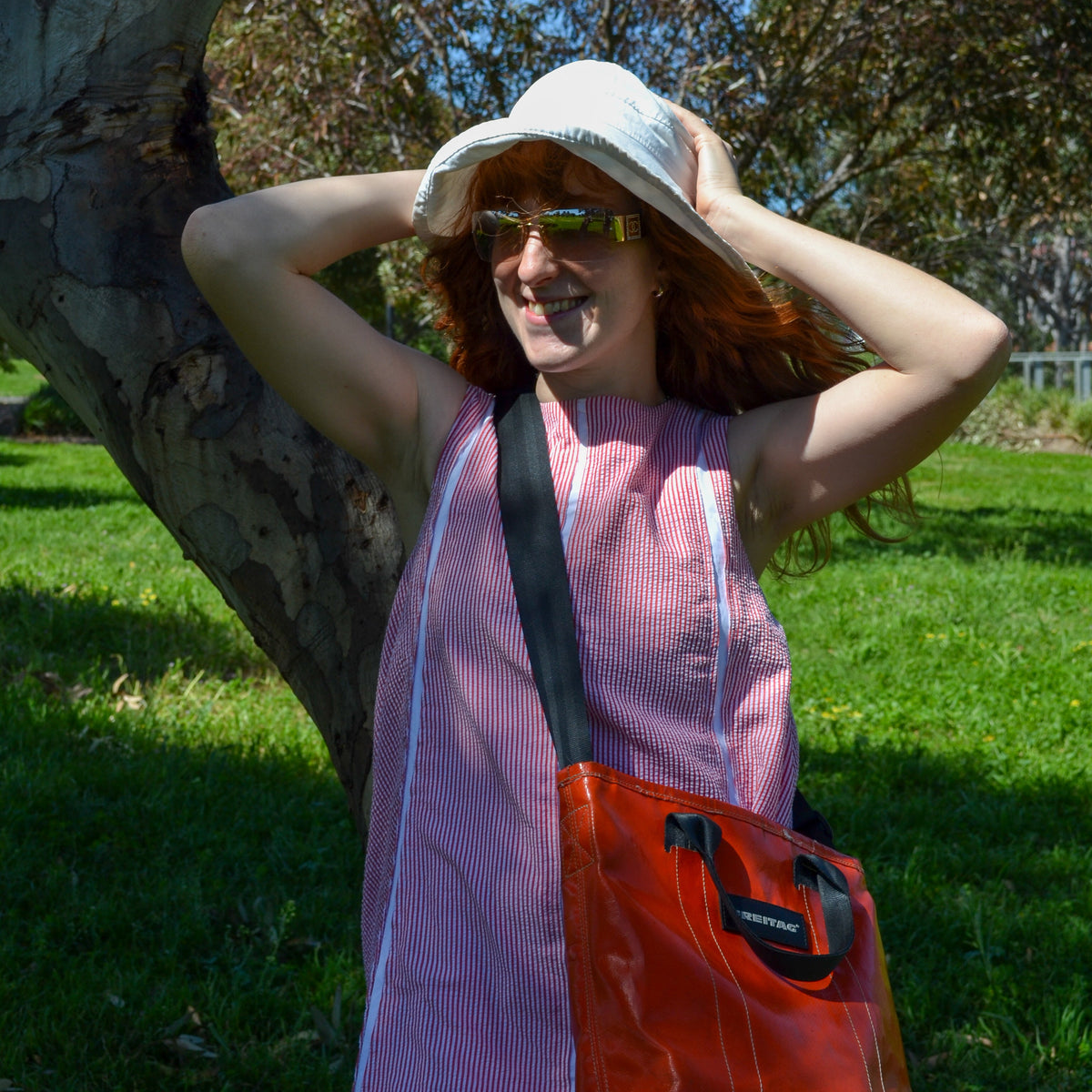 A woman outdoors wearing a white sun hat, green-tinted sunglasses, a pink sleeveless top, and carrying a bright orange-red shoulder bag with a black strap, standing near a tree trunk in a grassy park.