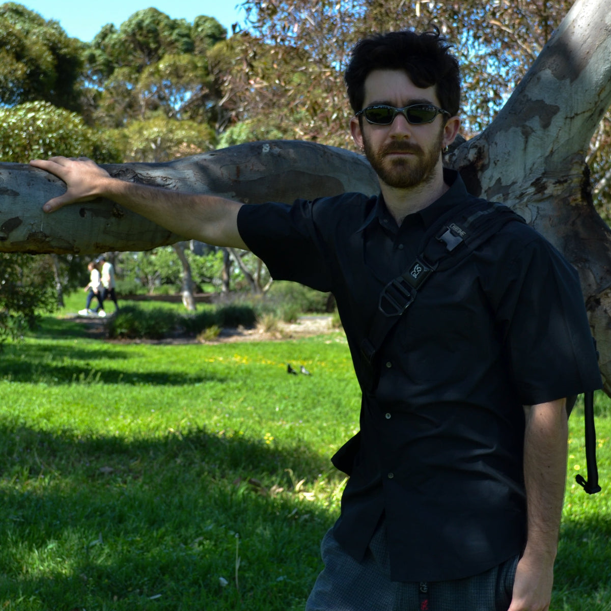 A man wearing a black short-sleeve button-up shirt, dark gray jeans, and sunglasses stands outdoors in a grassy park next to a large tree trunk, with his arm resting on it.