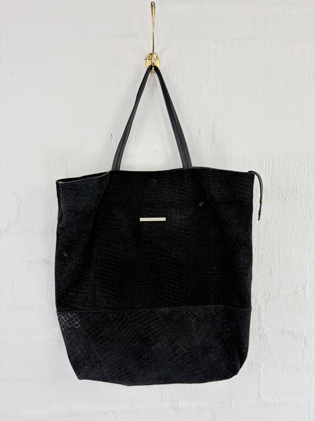 A black textured tote bag with black shoulder straps and a small silver rectangular accent on the front, hanging from a gold hook against a white wall.