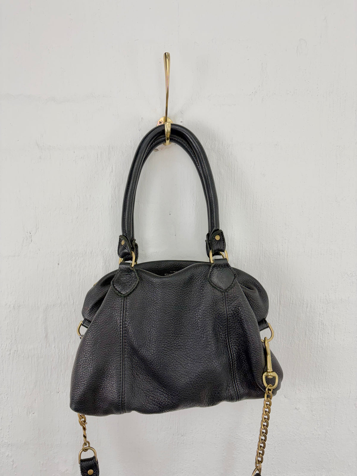 A black pebbled leather handbag with double top handles and gold-tone hardware, hanging from a gold wall hook against a white textured wall.