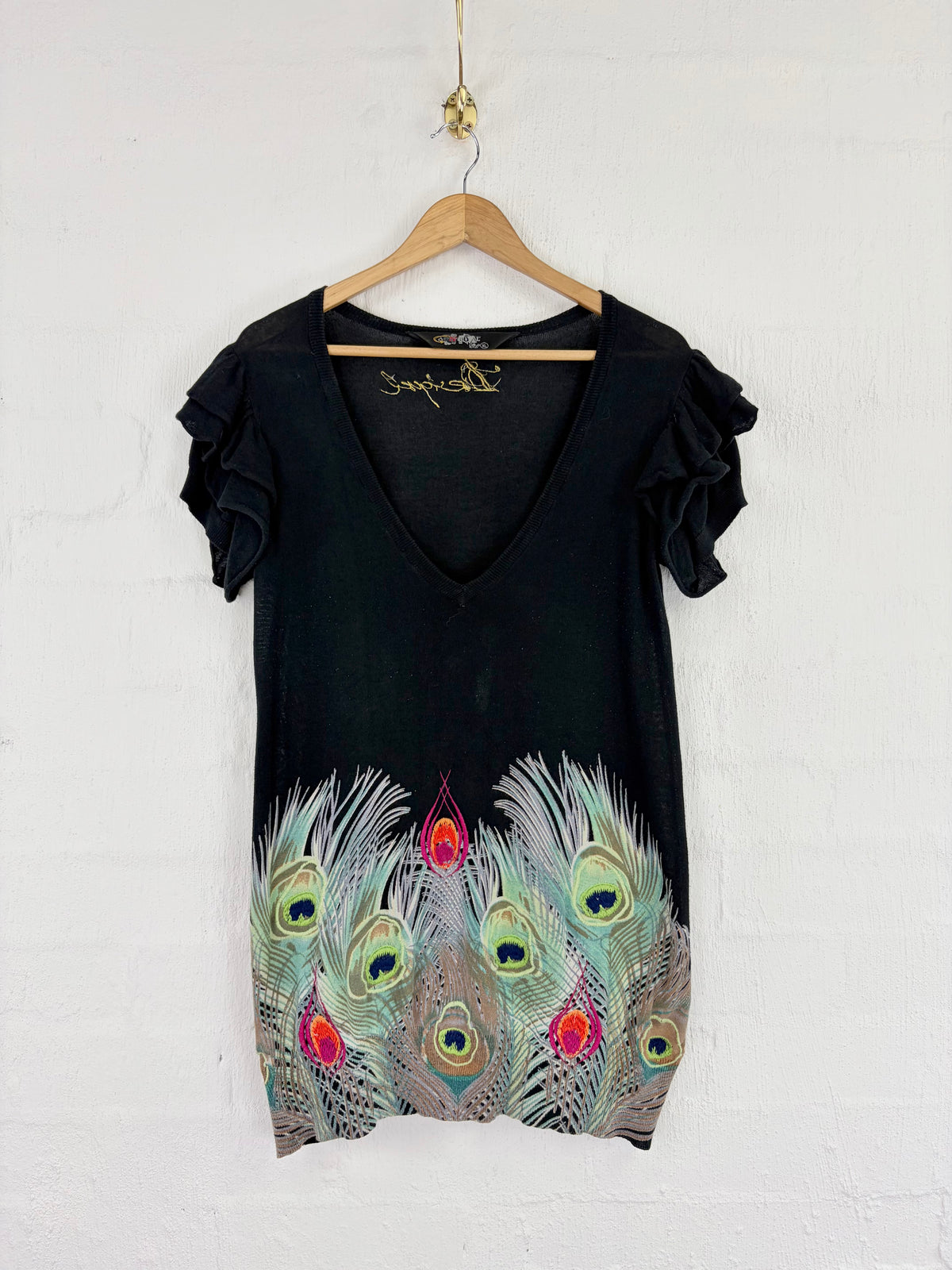 A black short-sleeve V-neck dress with ruffled sleeves and a colorful peacock feather print at the hem, hanging on a wooden hanger against a white brick wall.
