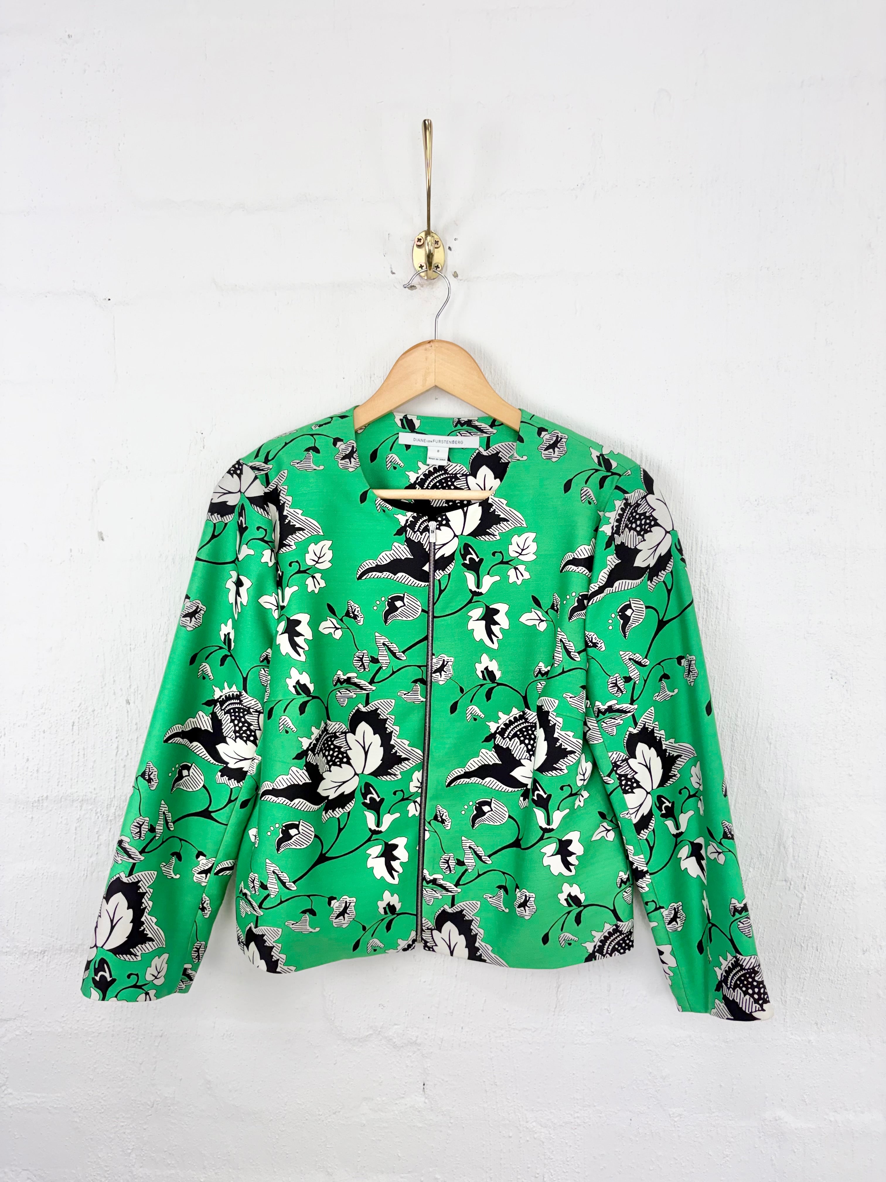 A green long-sleeve zip-up jacket with a black and white floral print, hanging on a wooden hanger against a white painted brick wall.