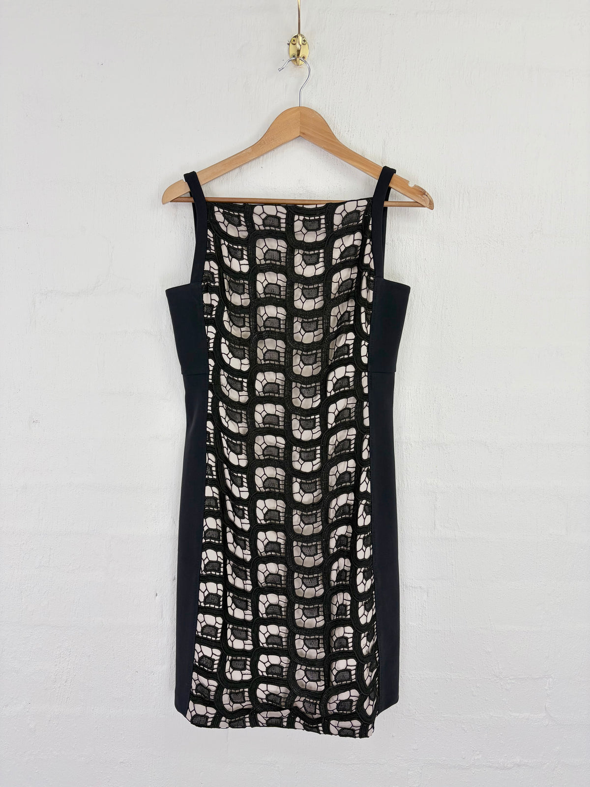 A sleeveless black and white patterned dress with solid black side panels, hanging on a wooden hanger against a white brick wall.
