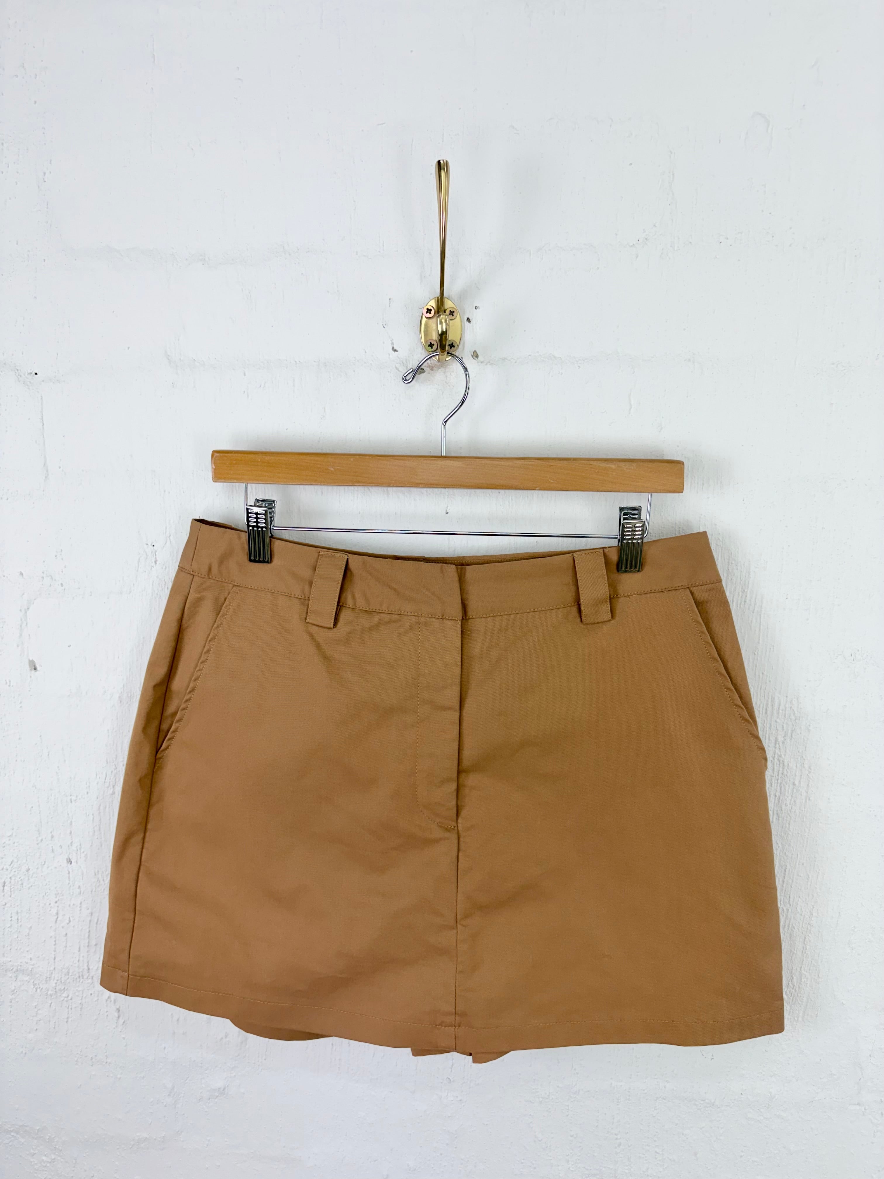 A tan mini skirt with belt loops and side pockets, hanging from a wooden hanger on a white brick wall.