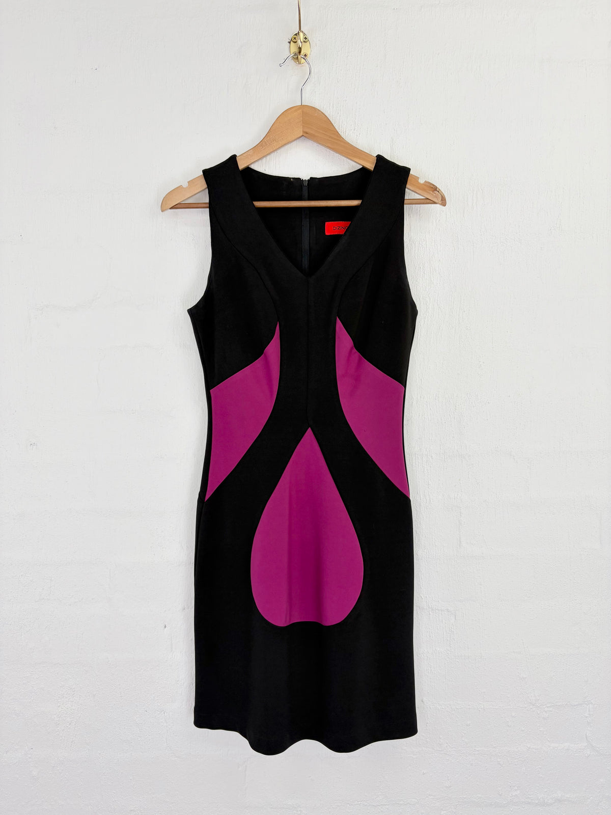 A sleeveless black dress with bold magenta color-blocked panels, hanging on a wooden hanger against a white painted brick wall.