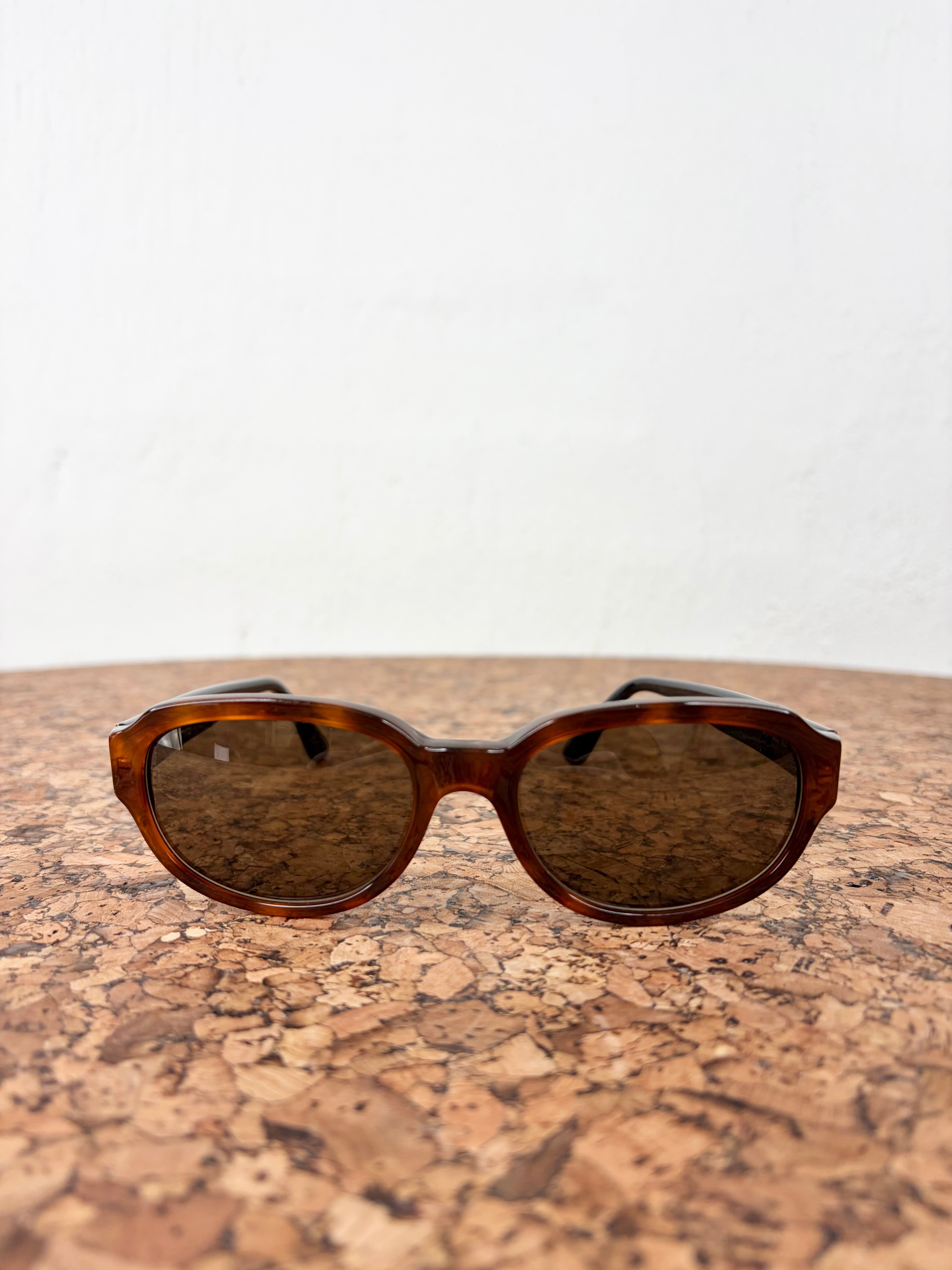 Oversized round sunglasses with a brown tortoiseshell acetate frame and dark brown lenses, resting on a cork surface against a white wall.