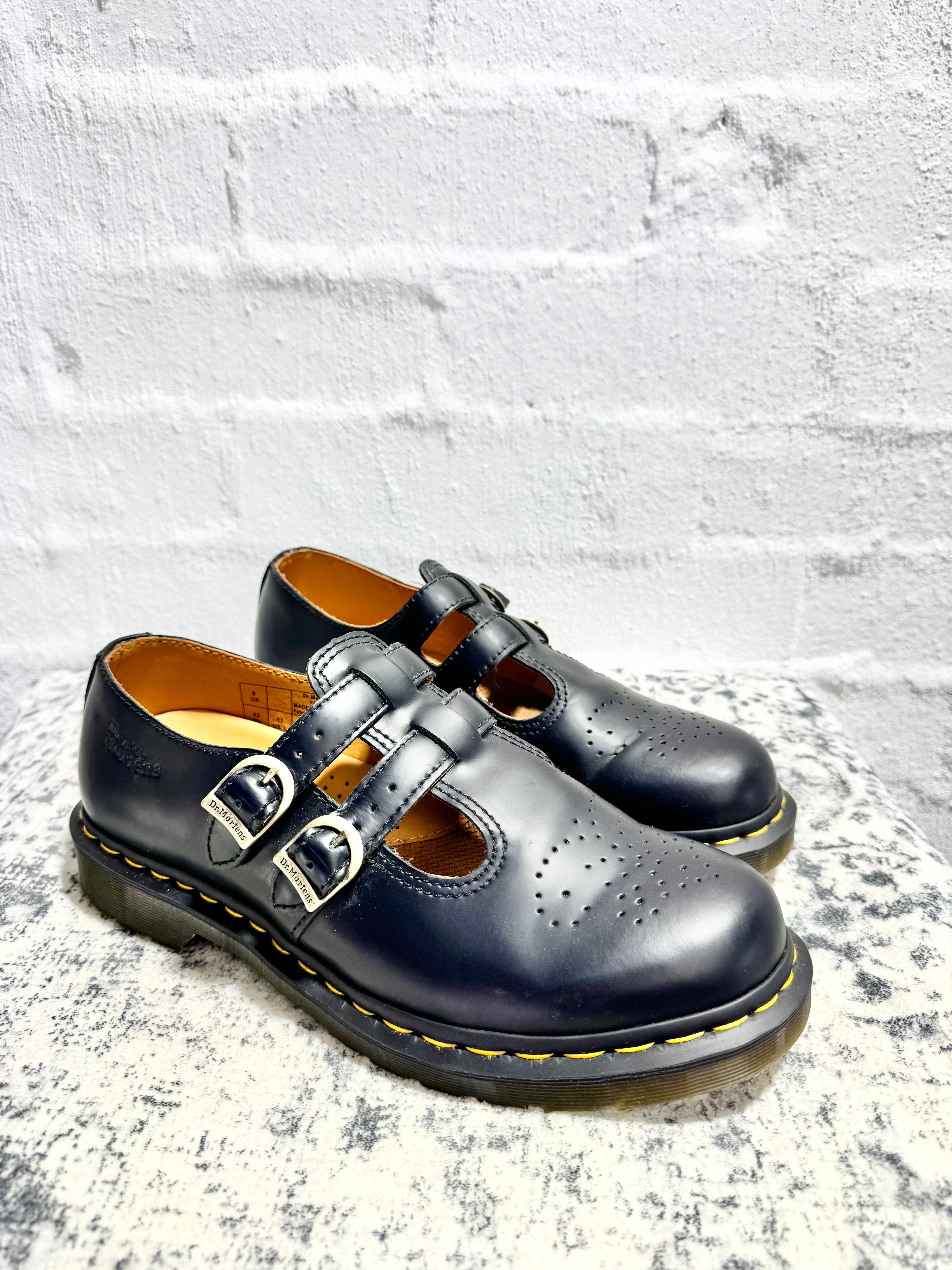 A pair of black leather Dr. Martens double strap Mary Jane shoes with silver buckles, displayed on a speckled stone surface against a white brick wall.
