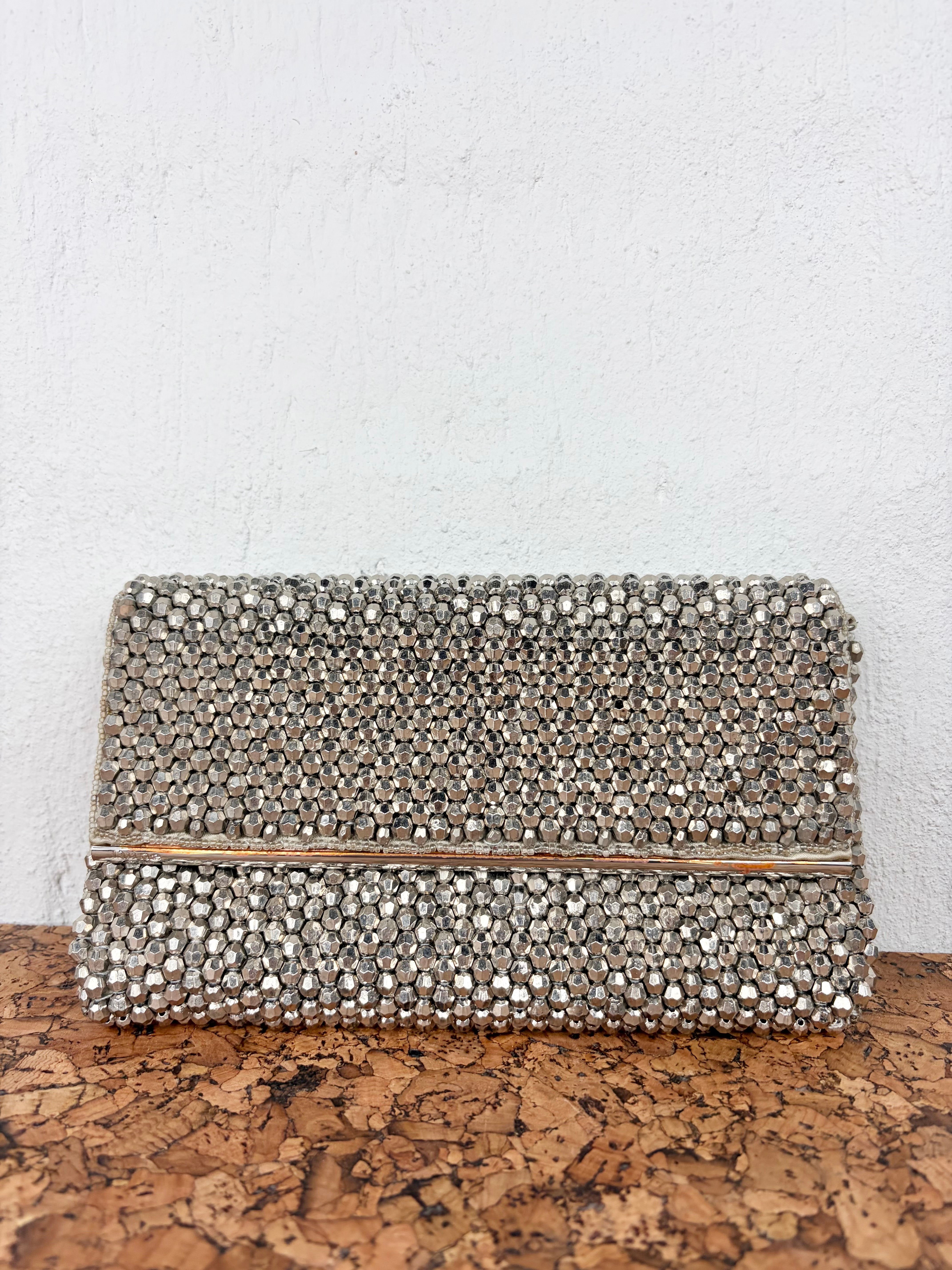 A rectangular clutch bag covered in silver rhinestones, featuring a thin metallic silver band across the center, resting on a cork surface against a white textured wall.